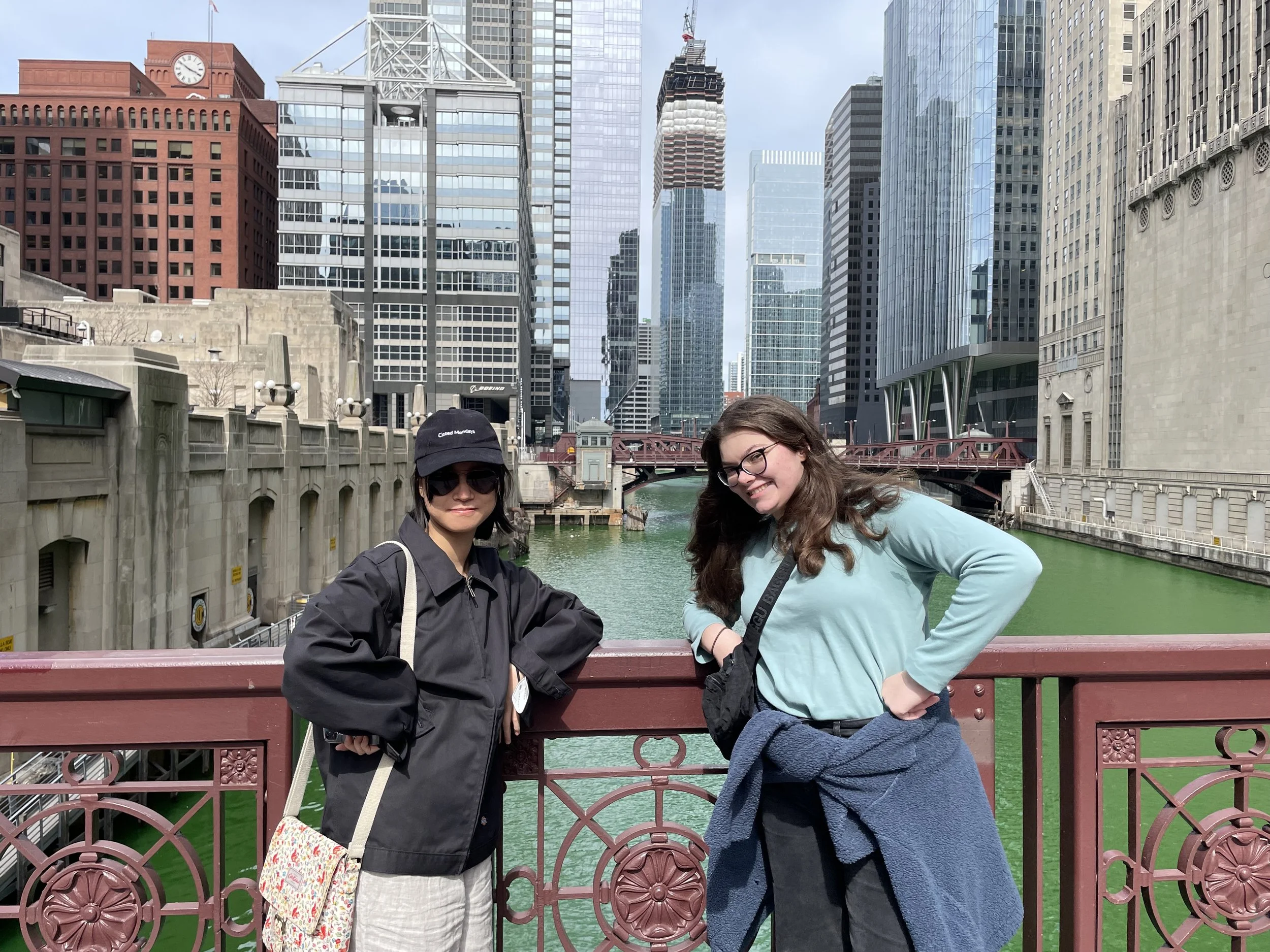 A young Asian person and a young white woman face the camera while leaning on a  reddish-brown painted ironwork bridge over the Chicago River, dyed a celebratory spring green.