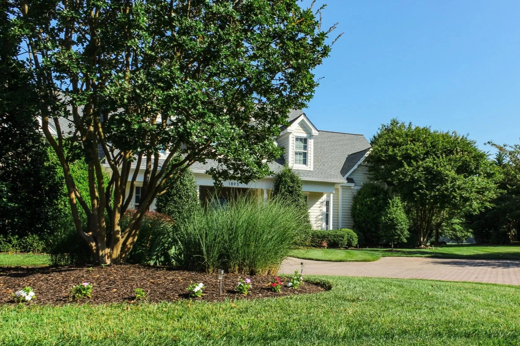 A well-maintained front yard of a suburban house with lush green grass, tall bushes, small trees, and a curved brick driveway. The house is white with a gray roof and has multiple windows, including a dormer window. Clear blue sky visible in the background.