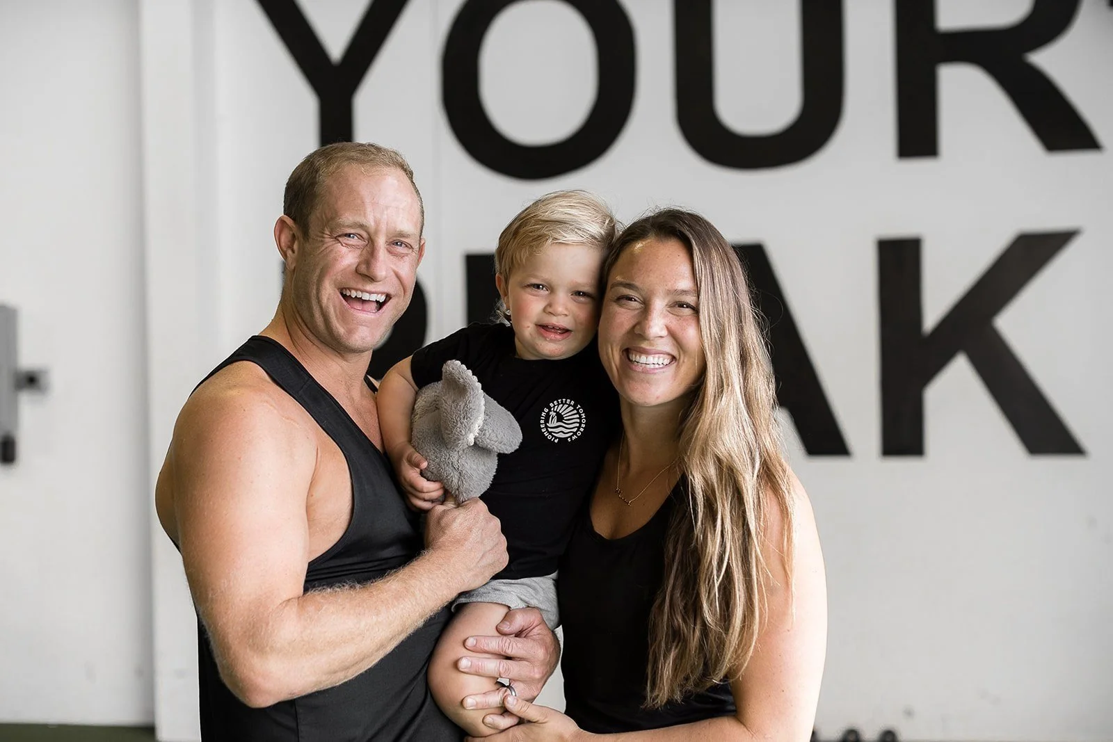 A family of three smiling indoors, with a man, woman, and a child holding a stuffed animal. Background features large partially visible letters on the wall.