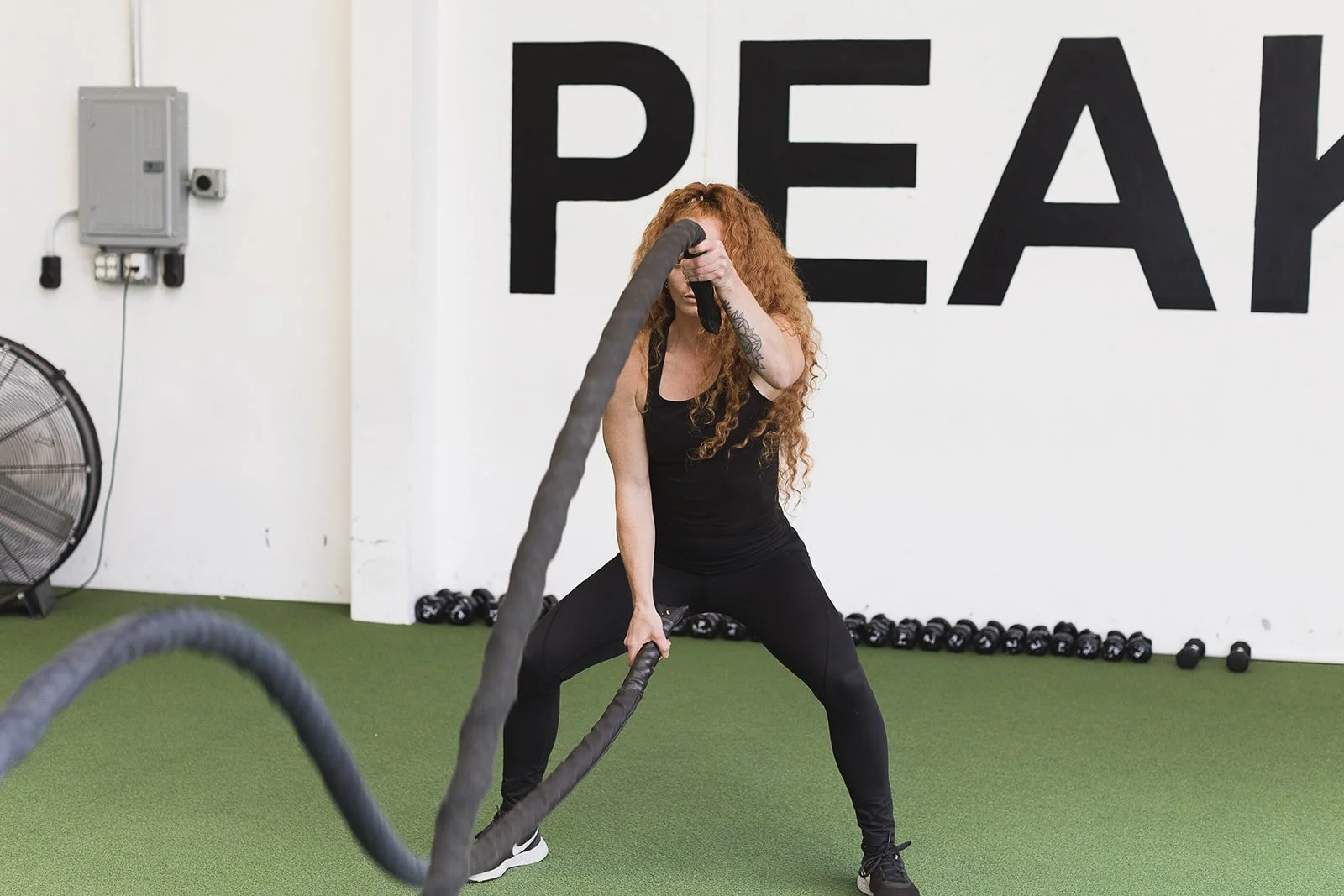 An Apex Fitness NW Trainer with long curly red hair in black athletic clothing doing battle ropes exercise in gym with large black 'peak' sign on white wall.  Photo taken at Apex Fitness NW, A Group Fitness Gym in Bellingham, WA.