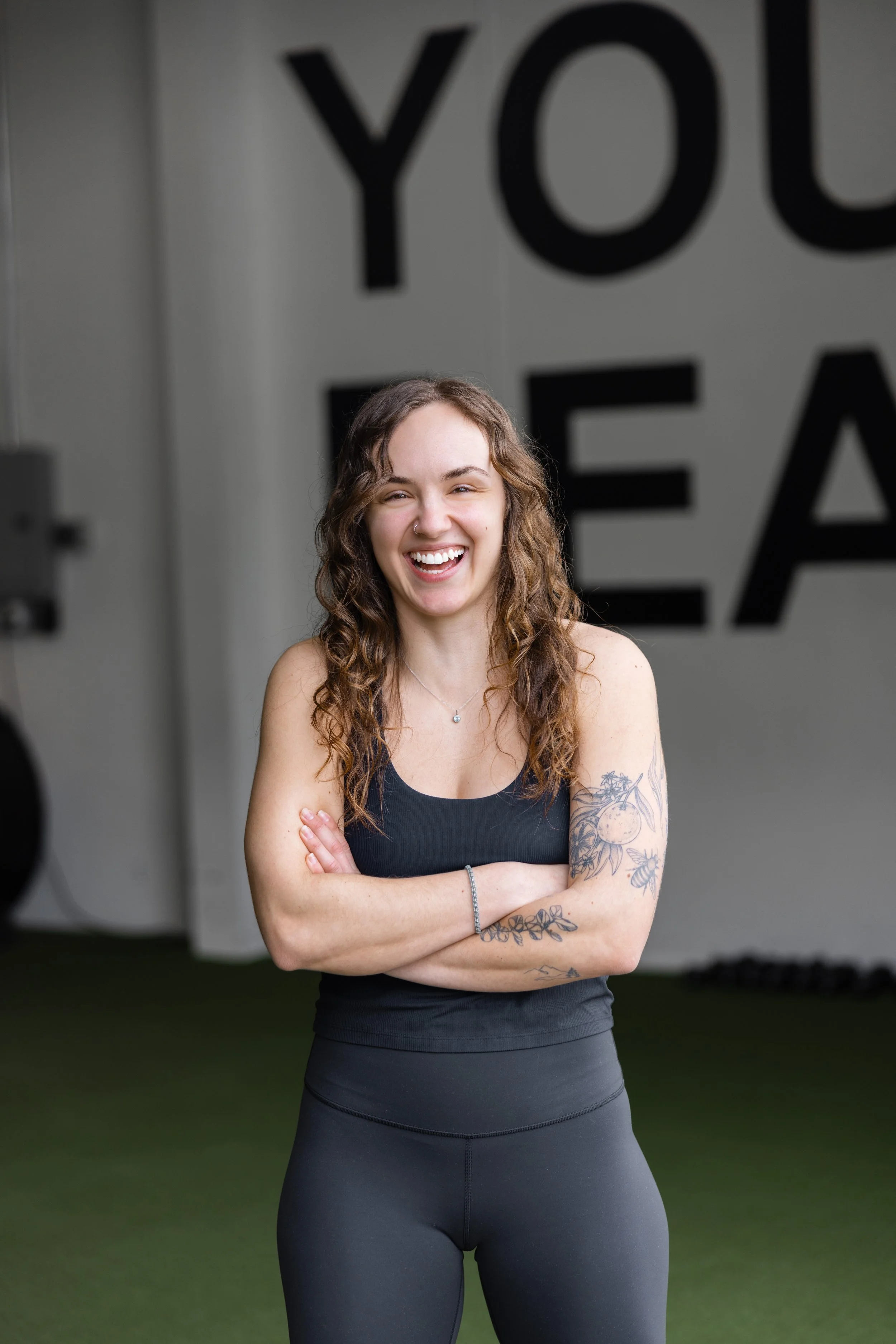 An Apex Fitness Trainer with curly brown hair, wearing a black athletic tank top and black leggings, standing with her arms crossed in a gym. She has tattoos on her arms and a nose piercing. (Apex Fitness NW, A Group Fitness Gym in Bellingham, WA)