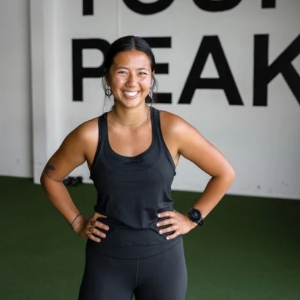 An Apex Trainer smiling workout attire standing with hands on hips in a gym, with a white wall and black text in the background that says 'Find Your Peak' at Apex Fitness NW, Group Training Gym.