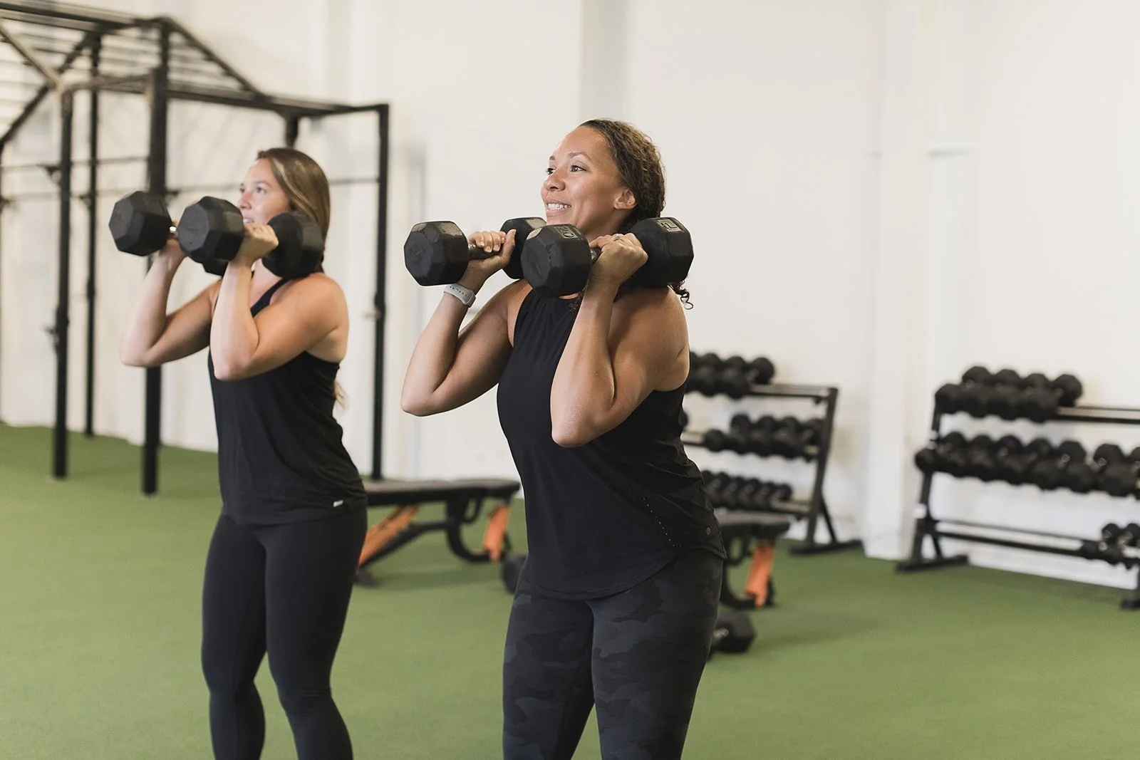 Two women lifting dumbbells in at Apex Fitness NW gym, in Bellingham, WA. They are wearing black workout clothes and standing on green turf. The background shows gym equipment and racks of dumbbells.