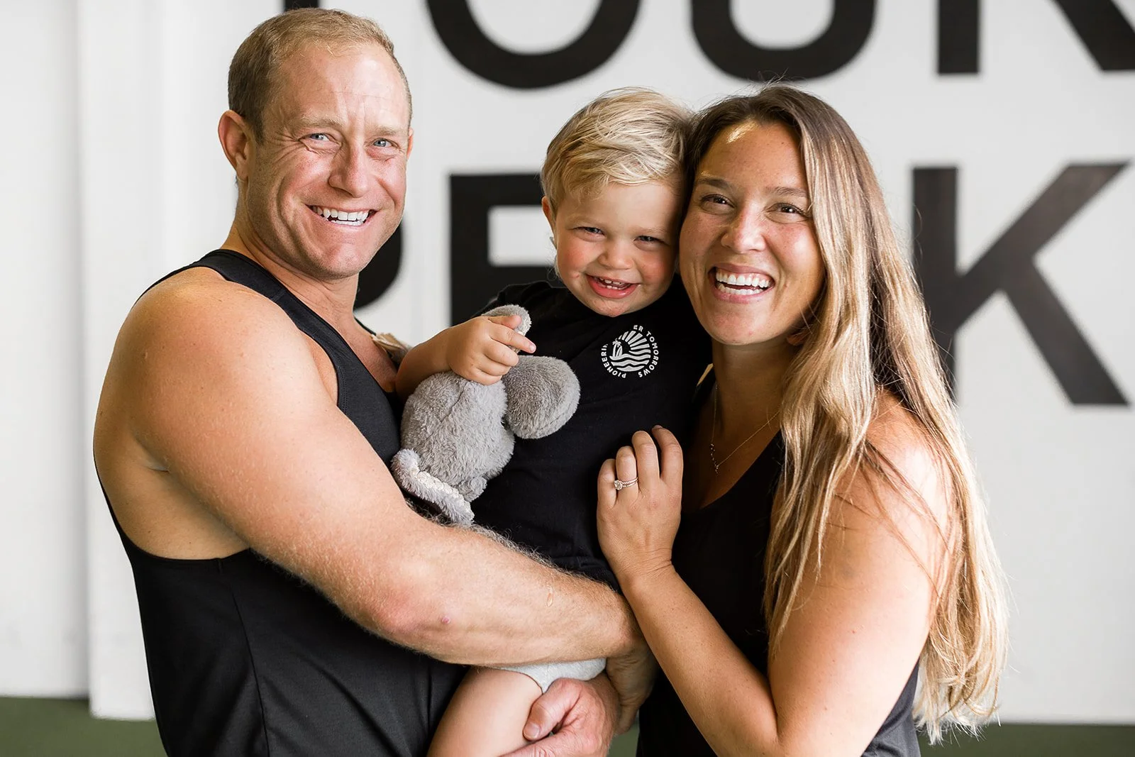 Smiling family with a child holding a stuffed toy in a gym setting.