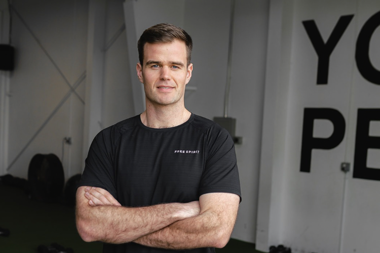An Apex Trainer with short dark hair and a slight smile, wearing a black t-shirt with a small logo that says 'Free Spirit', standing with arms crossed in a Apex Fitness NW fitness studio. (Apex Fitness NW, Gym in Bellingham, WA)