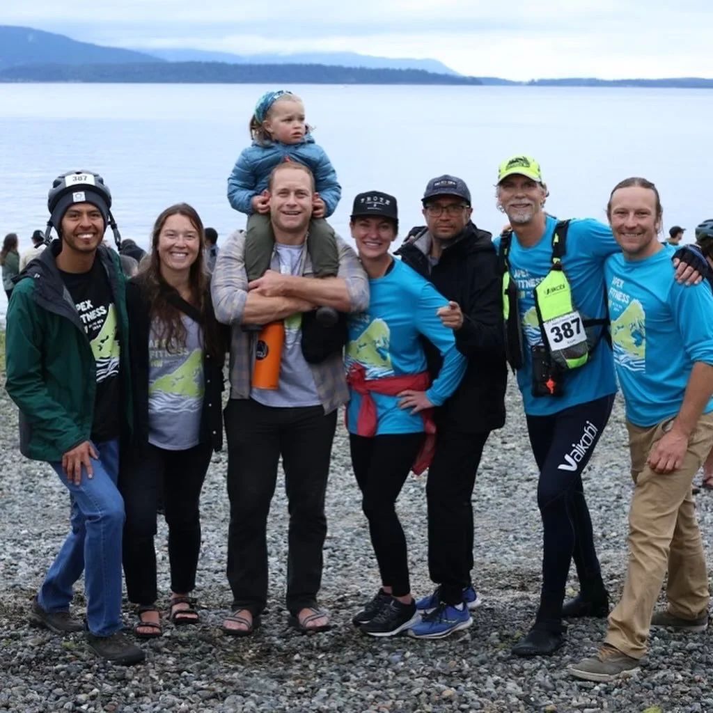 Apex Fitness NW Group standing on a pebble beach with water and mountains in the background, smiling for a photo, some wearing running gear and bib numbers, one person carries a child on his shoulders.