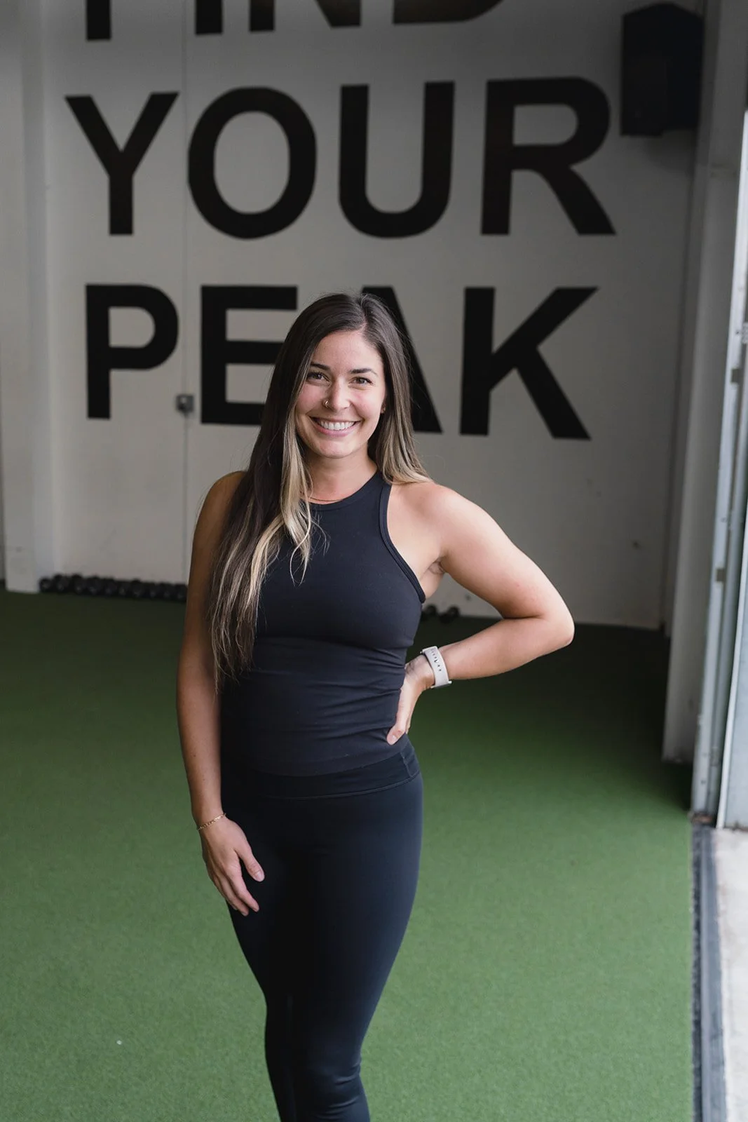 Woman in black workout attire standing on green turf, smiling, with "Find Your Peak" in large letters on the wall behind her.
