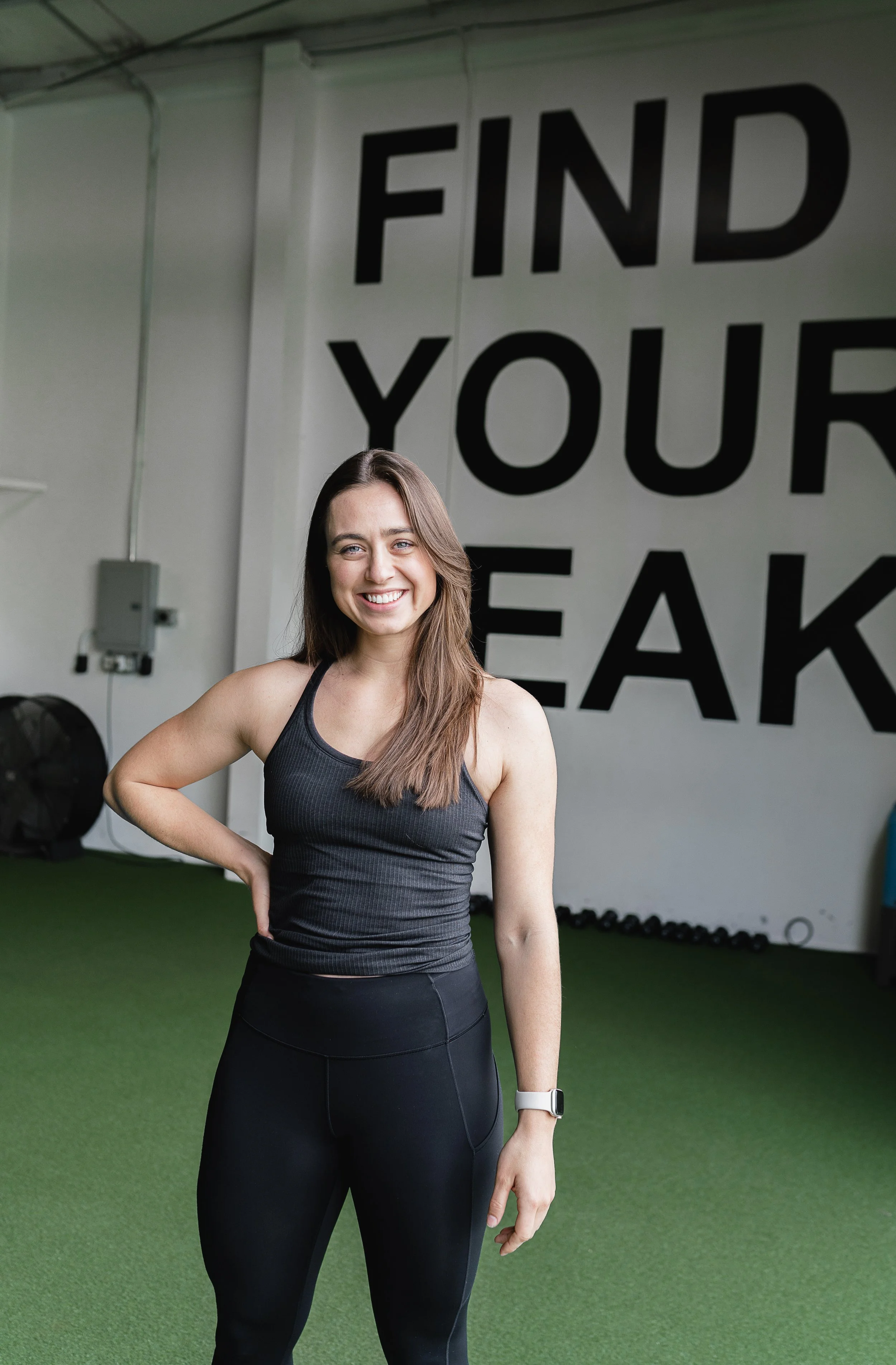An Apex Trainer smiling in workout attire standing in at the Apex gym with a motivational sign that says 'FIND YOUR PEAK' on the wall behind her. (Apex Fitness NW, Gym in Bellingham, WA)