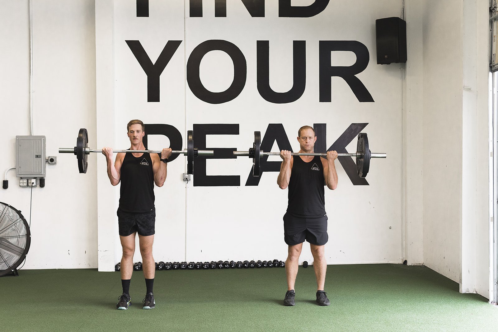 Two men standing side by side lifting barbells at Apex Fitness NW Gym. The background has a large sign that reads 'FIND YOUR PEAK'. Both men are dressed in black workout attire.  Photo taken at Apex Fitness NW, A Group Fitness Gym in Bellingham, WA.