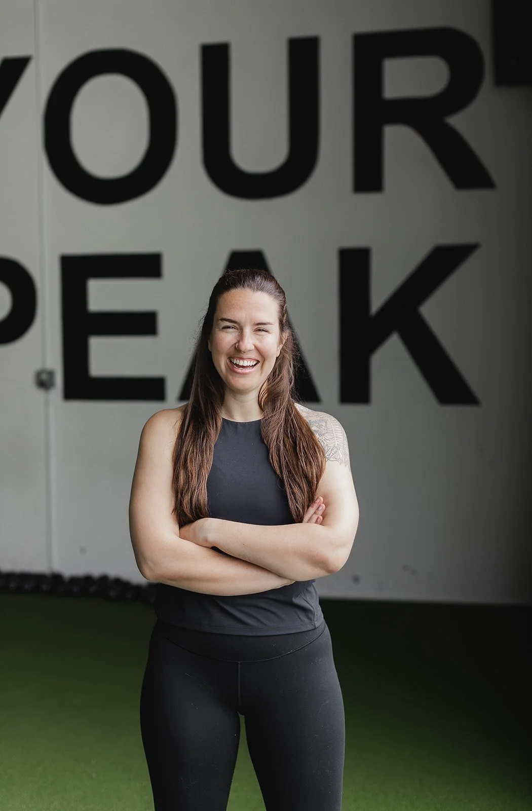 An Apex Group Fitness Trainer with long brown hair, wearing a black athletic tank top and black leggings, standing with her arms crossed and smiling in front of a wall with black lettering that says "Find Your Peak."