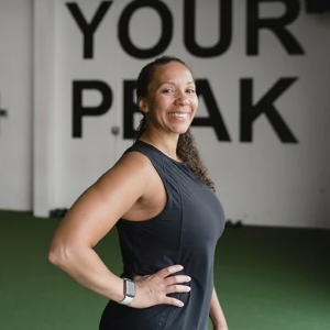 An Apex Trainer smiling with curly hair at Apex Fitness NW Gym in Bellingham, standing in front of a wall with large black text that reads 'Find Your Peak'. (Apex Fitness NW, Gym in Bellingham, WA)