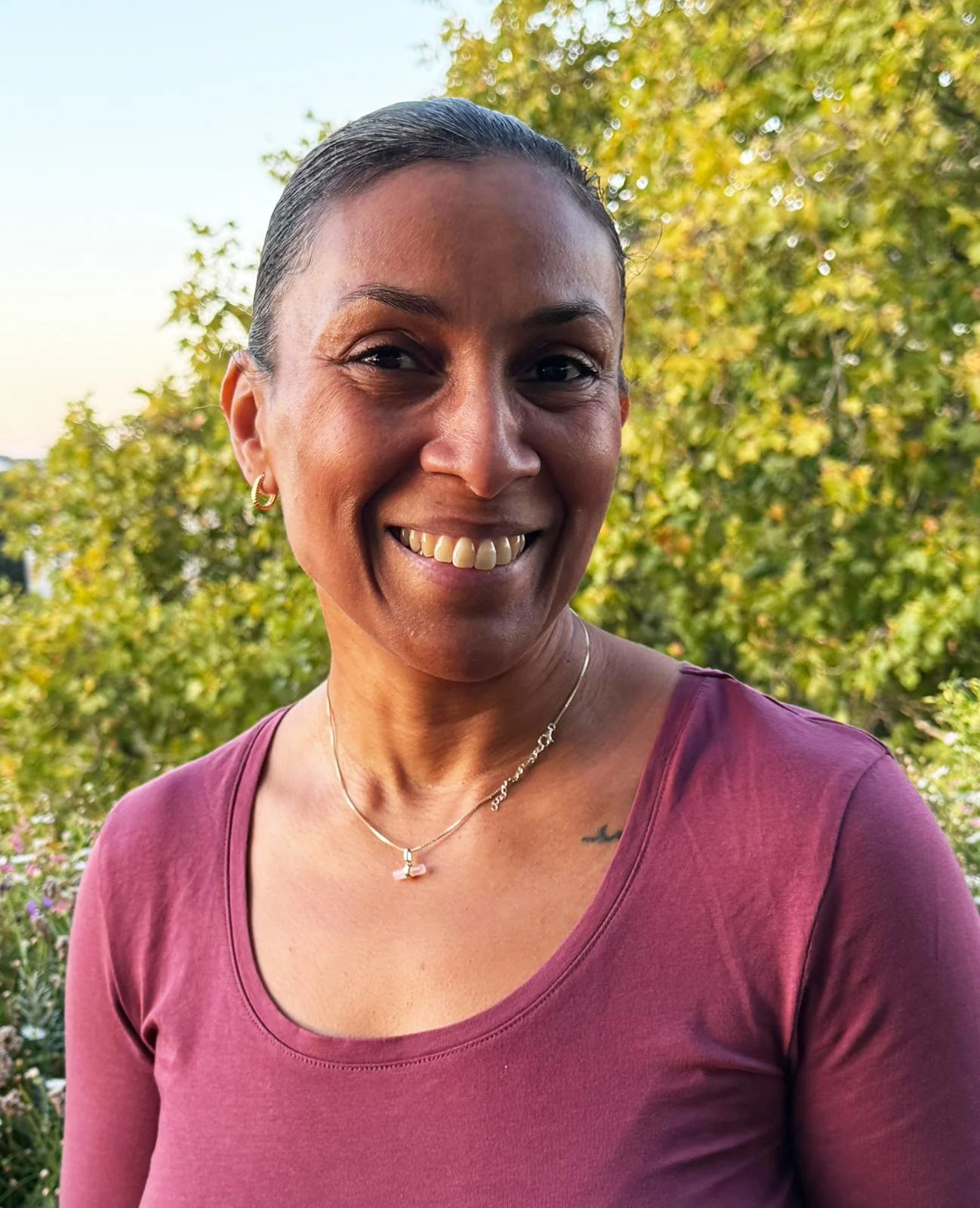 A woman smiling outdoors with green trees and a clear sky in the background, wearing a maroon top and jewelry.
