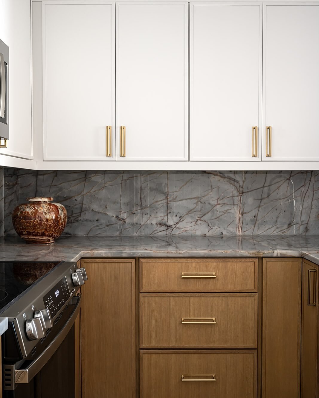 This kitchen features a beautiful, modern design with a striking quartzite backsplash and countertop that includes dramatic veining. The space is complemented by sleek white upper cabinets, warm wood lower cabinets, and brushed gold hardware, creatin
