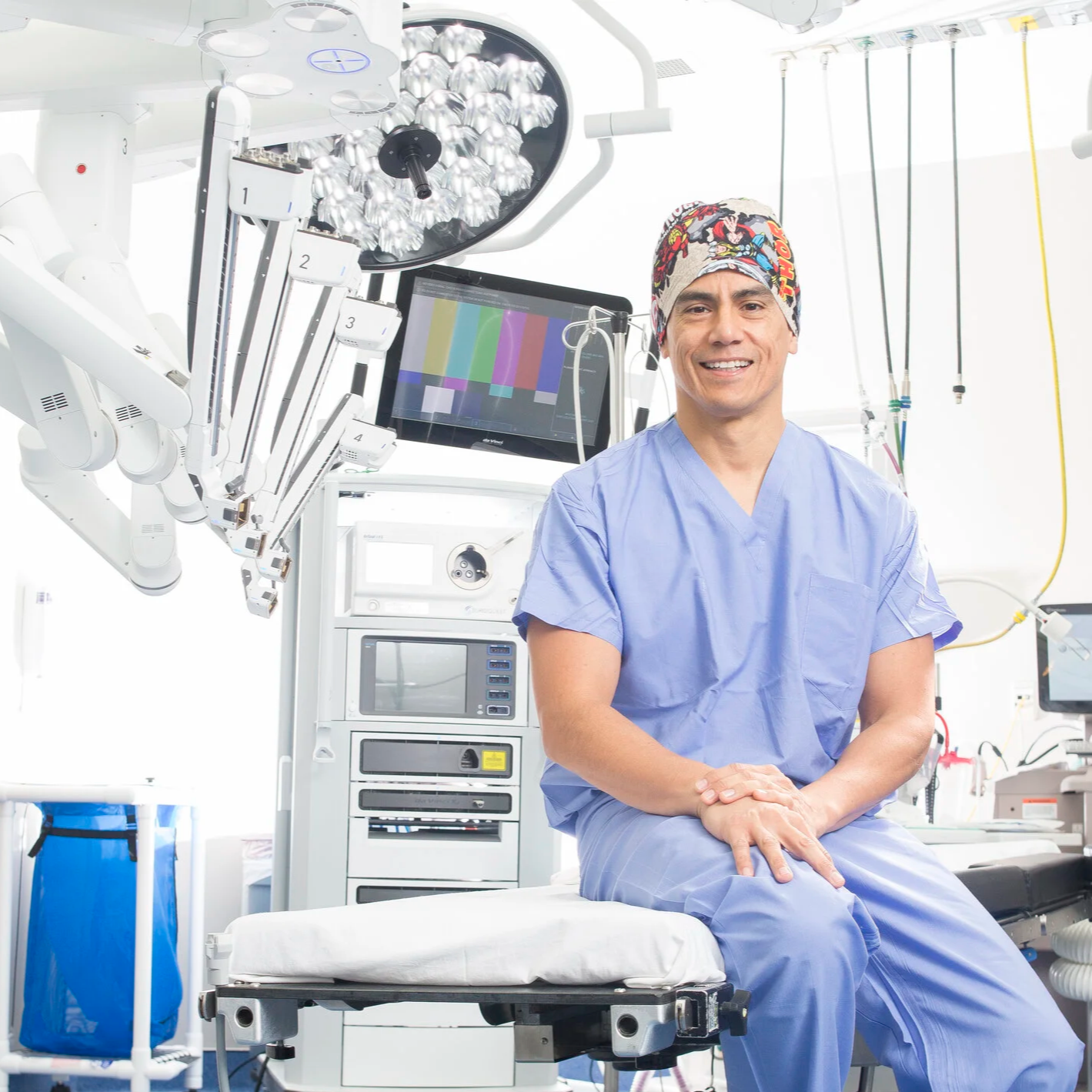 A male healthcare professional sitting in a hospital room, wearing blue scrubs and a colorful cap, surrounded by medical equipment and monitors.