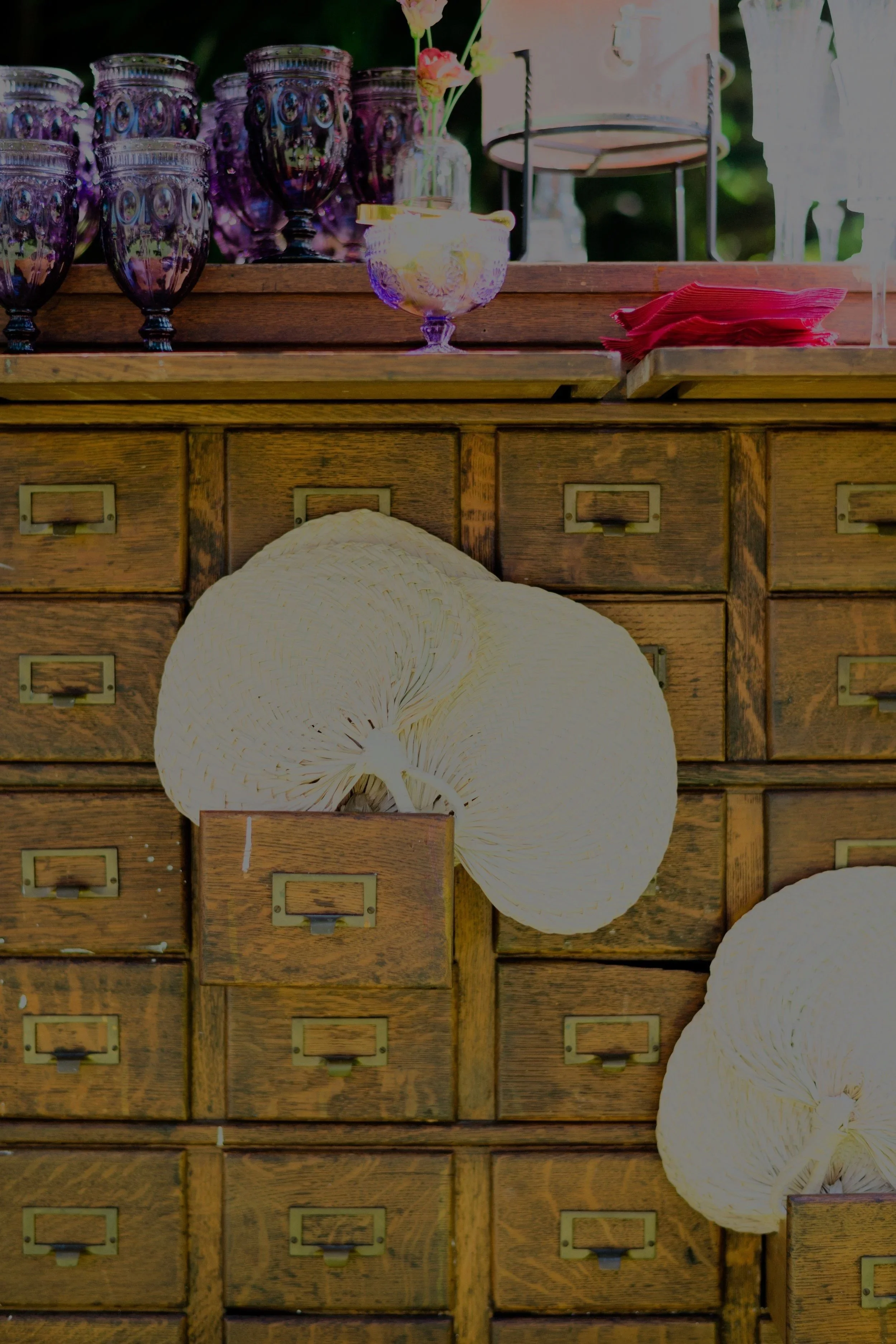 Vintage wooden filing cabinet with multiple small drawers, decorated with white woven hats hanging from the drawers, and purple glassware and a cake on top.