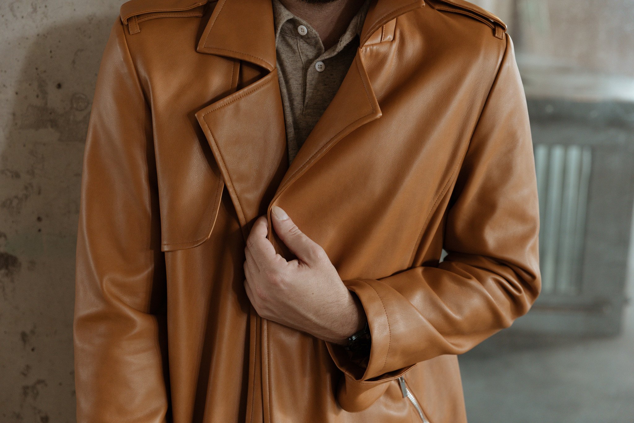 Person holding a brown leather jacket in front of a concrete wall.