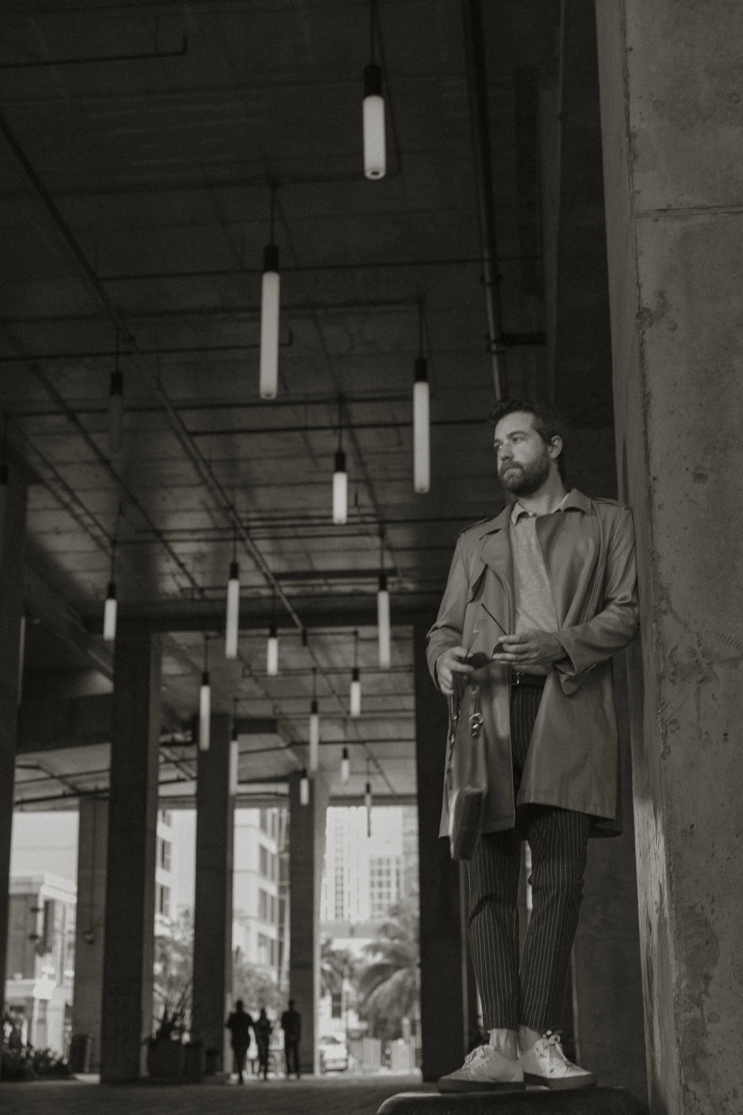 A man leaning against a concrete wall in an urban parking garage, holding a phone and a bag, with tall buildings visible in the background.