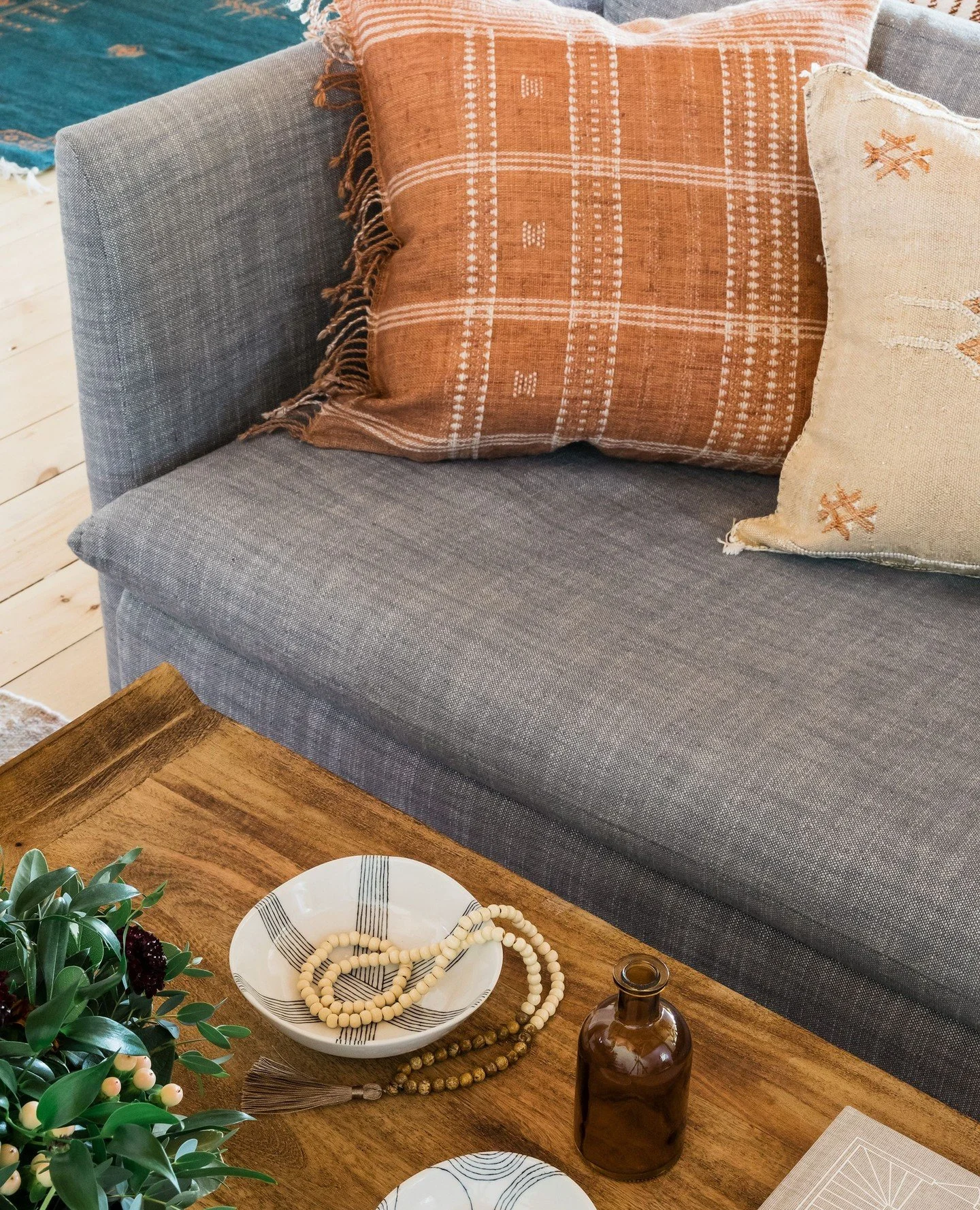 A rust-toned handwoven pillow with fringe edge and an embroidered natural linen against a grey linen sofa. A teal kilim rug behind. A ceramic dish, bead strand, and amber bottle on the mango wood coffee table. In an open barn interior shaped entirely