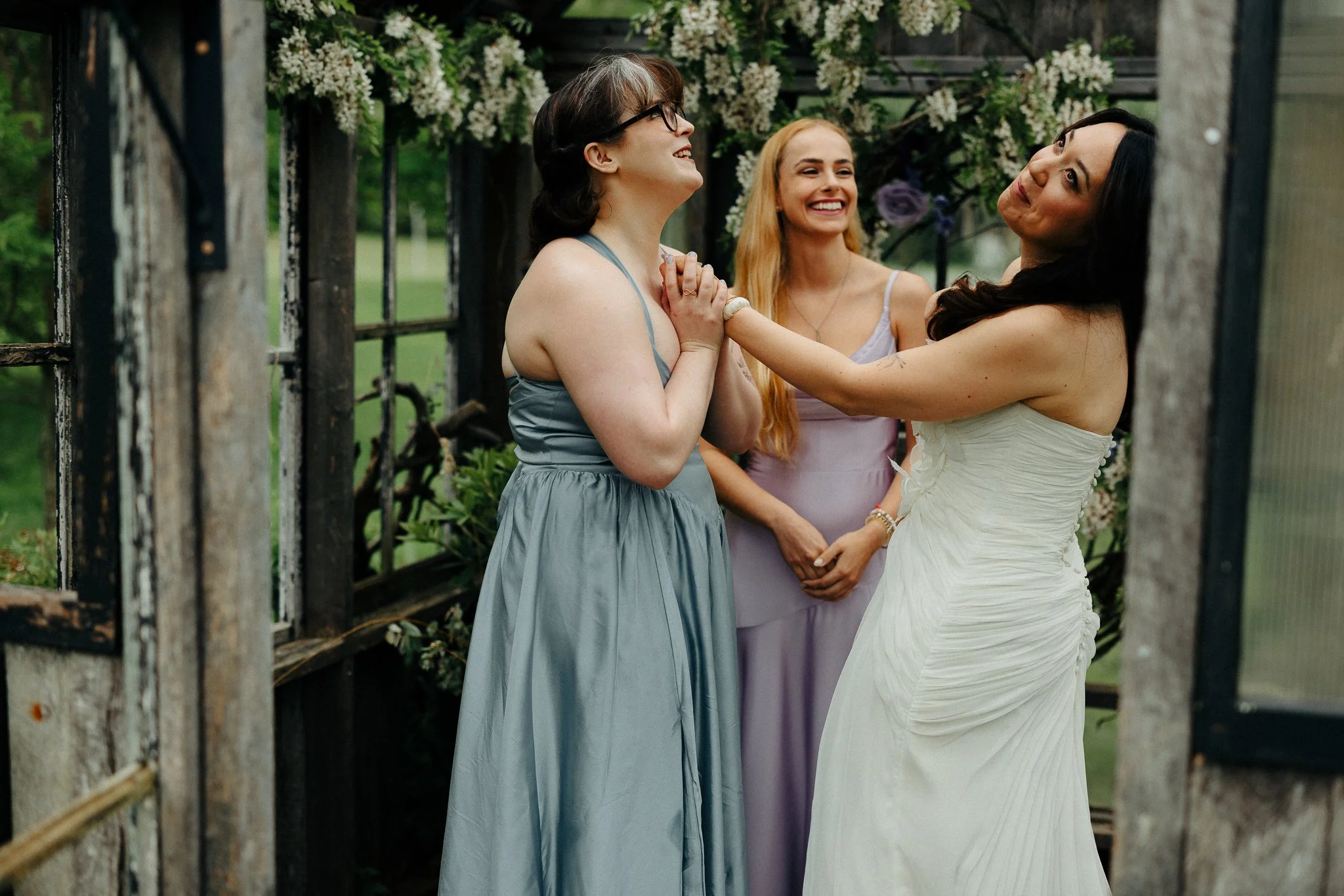 Three women at a wedding ceremony, with one woman in a white wedding dress and two women in pastel-colored dresses, smiling and holding hands in an outdoor garden setting.