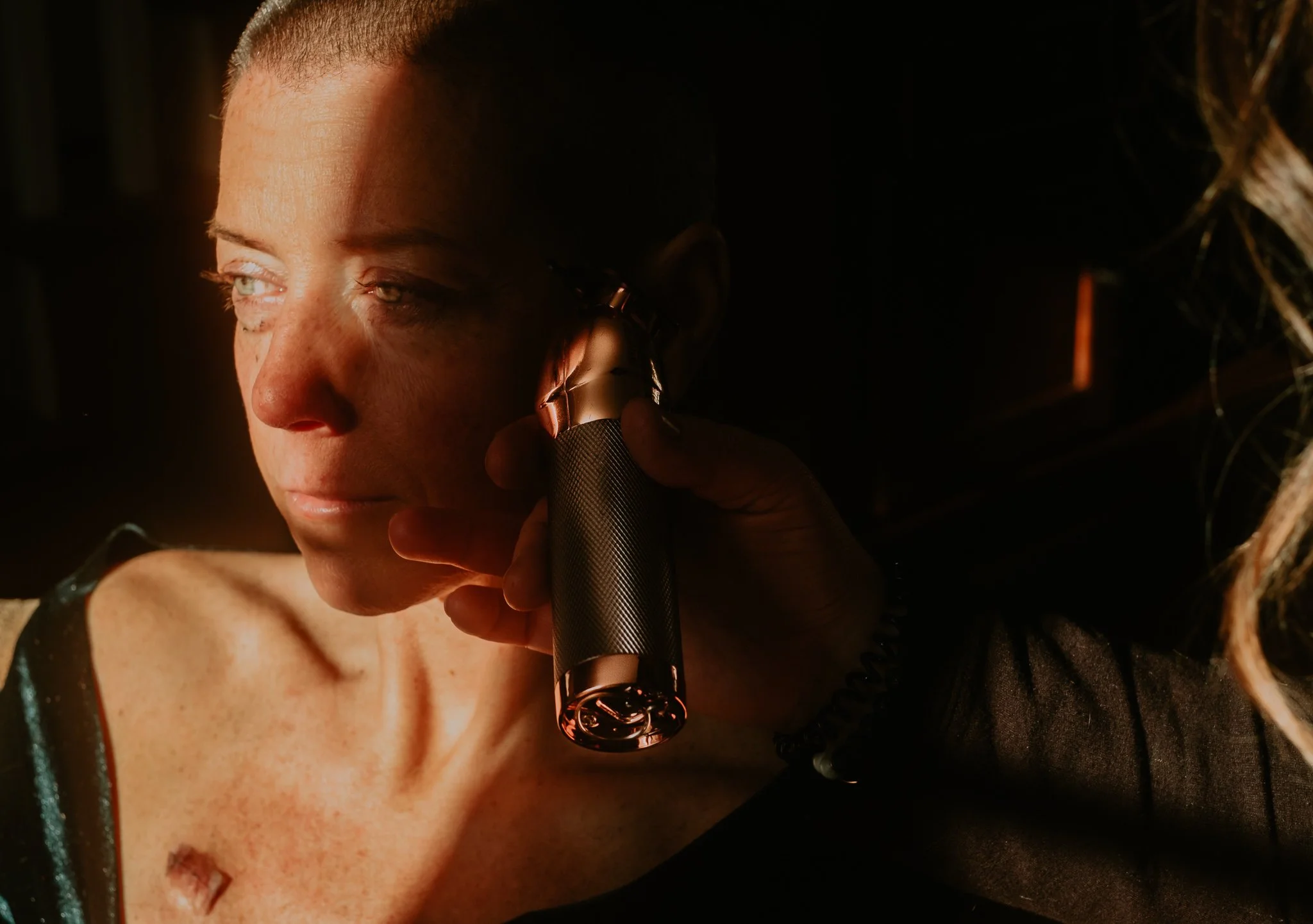 A woman with short hair and light skin sitting in dark lighting, having her ear examined or cleaned with a medical device used by a person whose hand is visible.