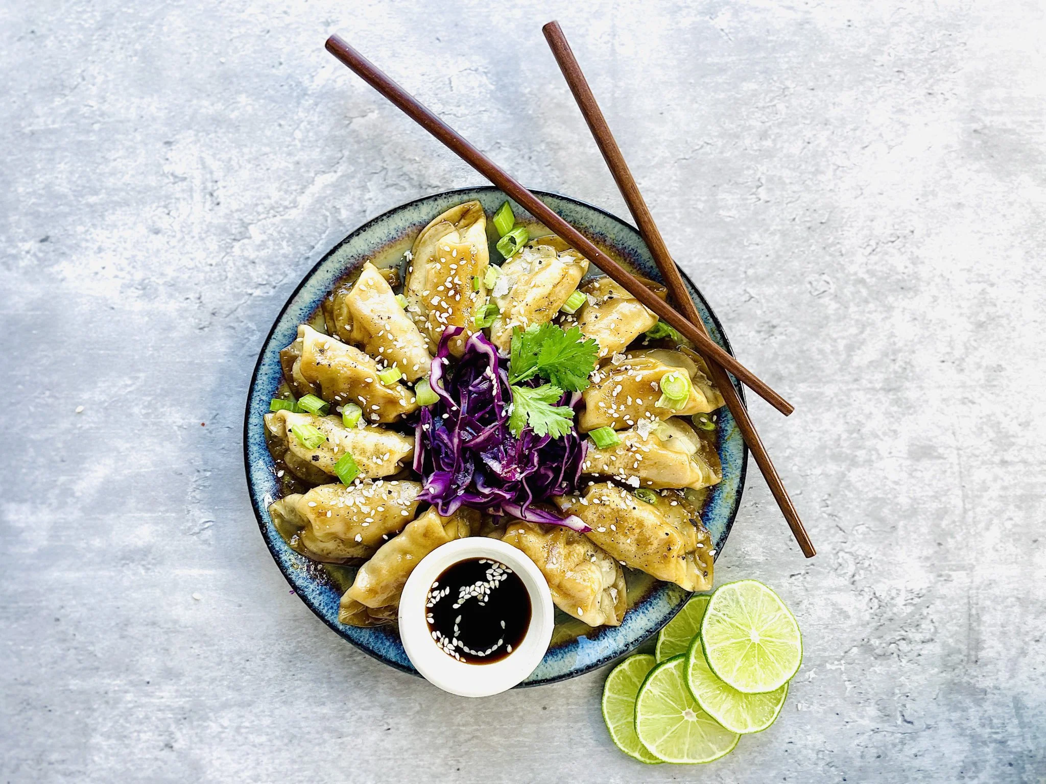 A bowl of dumplings garnished with green onions, sesame seeds, purple cabbage, cilantro, accompanied by a small dish of soy sauce, and slices of lime on the side.