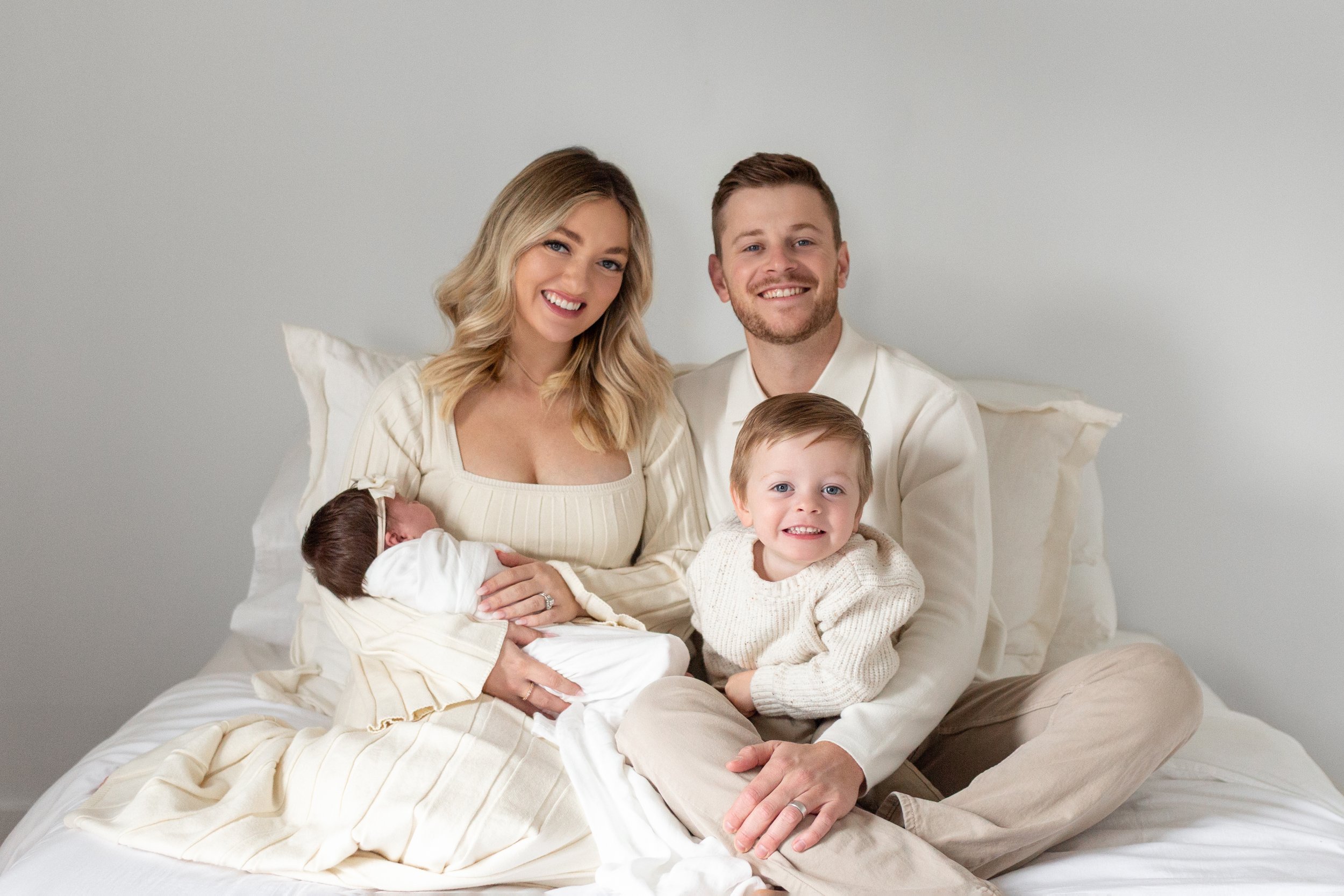 A family of four sitting on a bed, smiling and posing for a photo. The mother holds a newborn, the father sits beside her, and an older toddler sits on his lap. All are dressed in cream-colored clothing, with a plain white wall background in studio.a