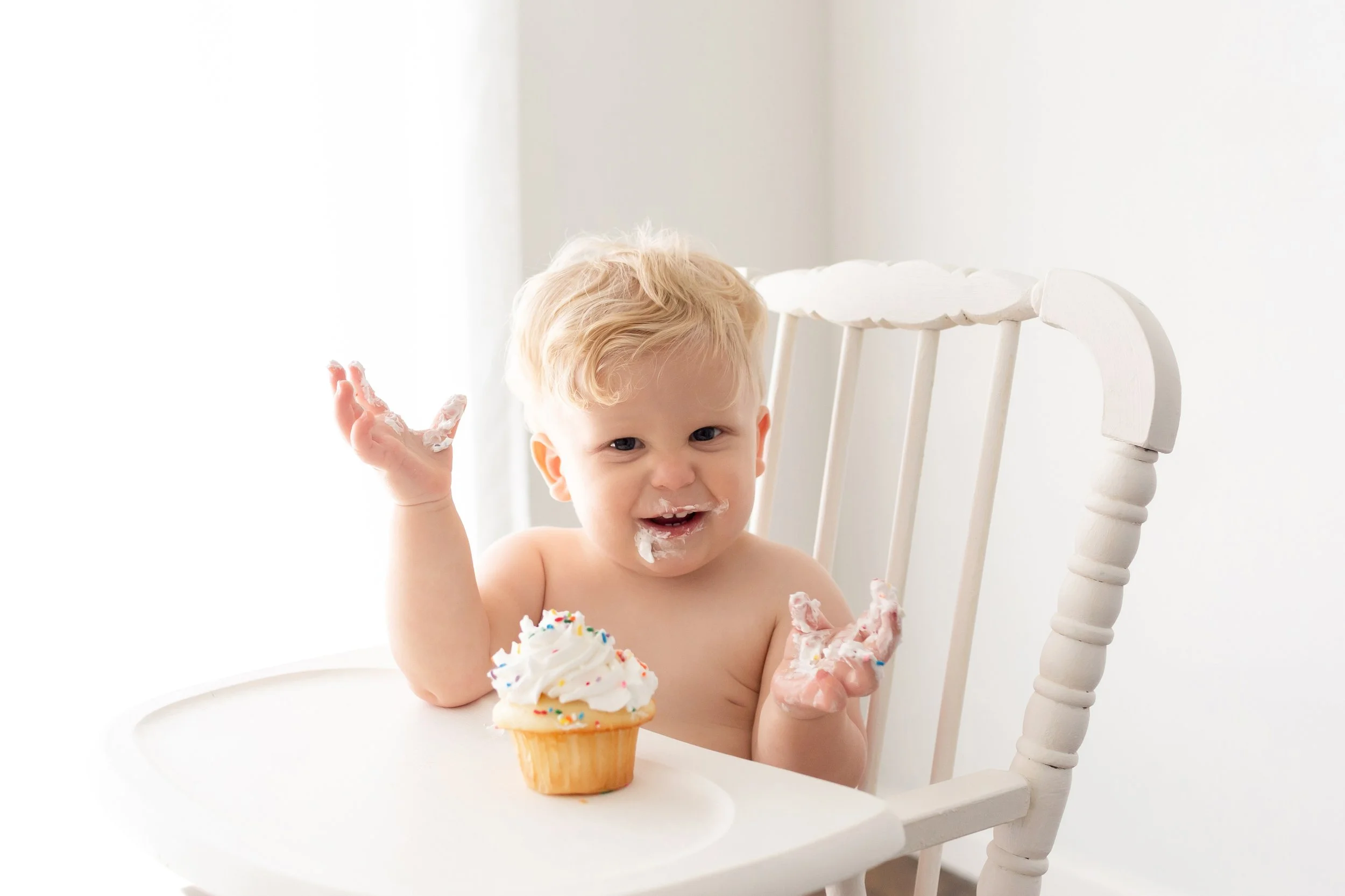 A smiling baby with messy hands and face sits in a high chair, enjoying a cupcake with white frosting and colorful sprinkles in a Fayetteville, Arkansas studio..