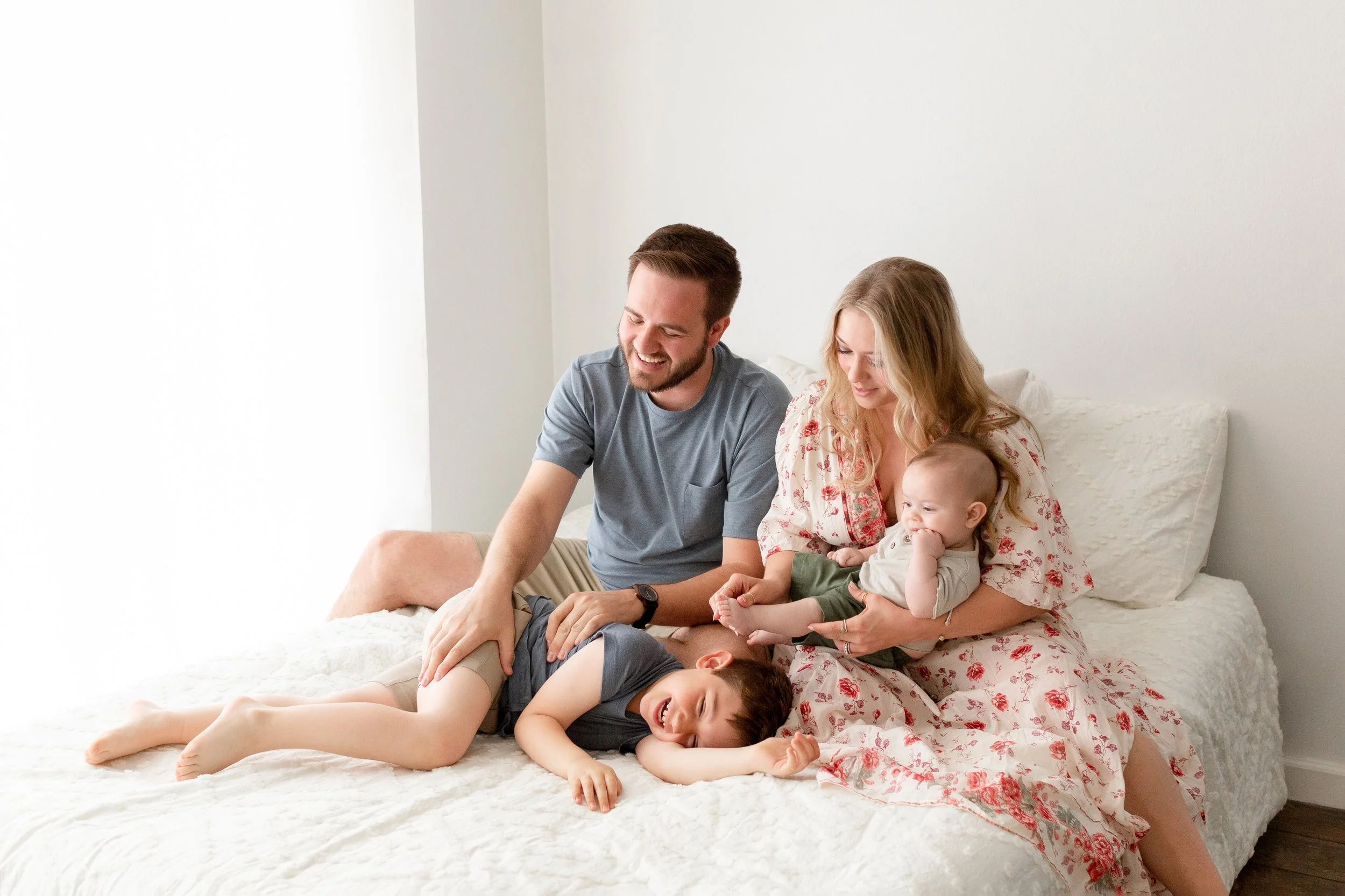 A family of four sits on a bed, laughing and playing together in a bright, cozy bedroom in a Fayetteville, Arkansas studio..