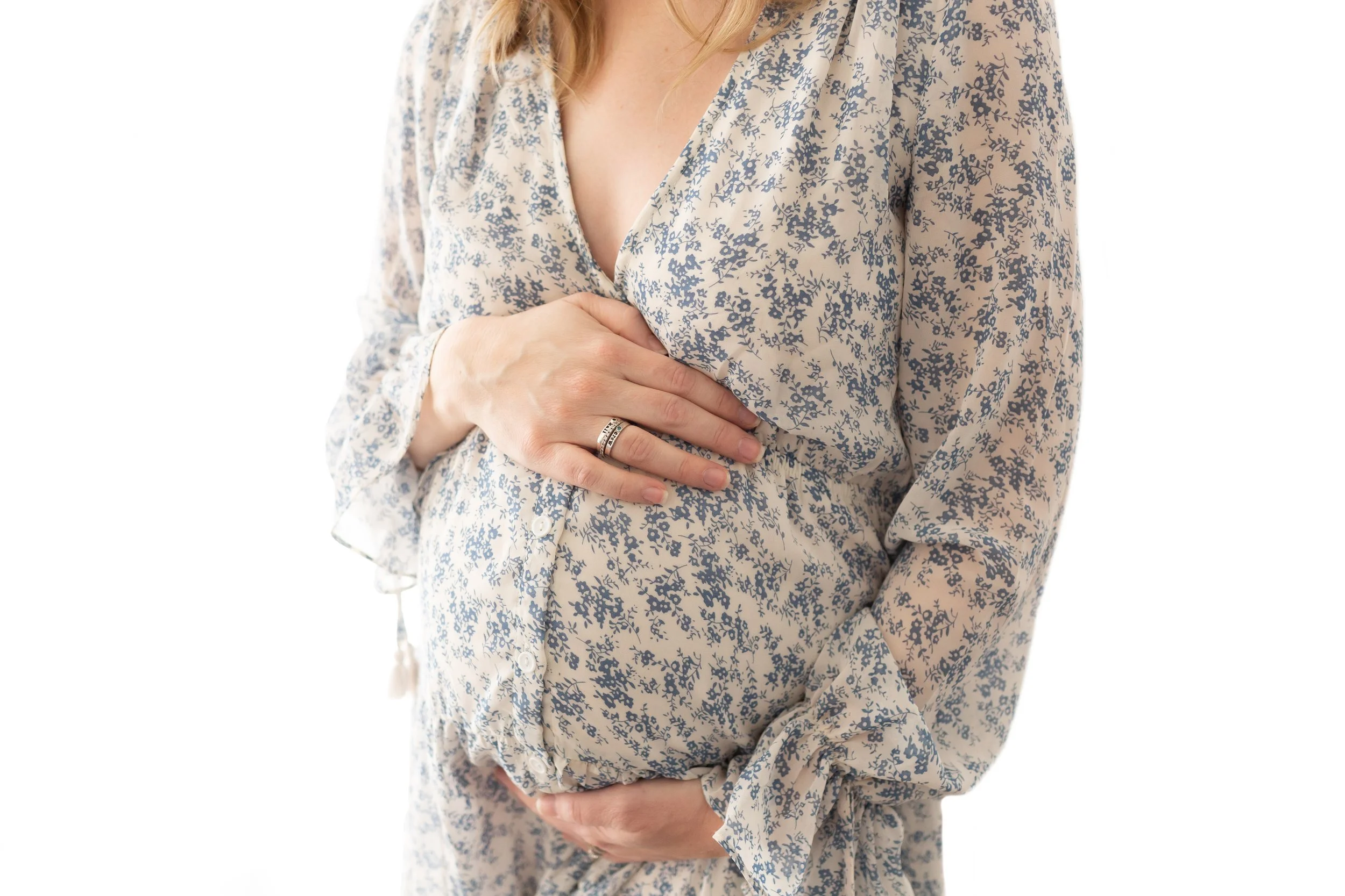 A pregnant woman in a floral dress cradles her belly with both hands against a white background in a Fayetteville, Arkansas studio.
