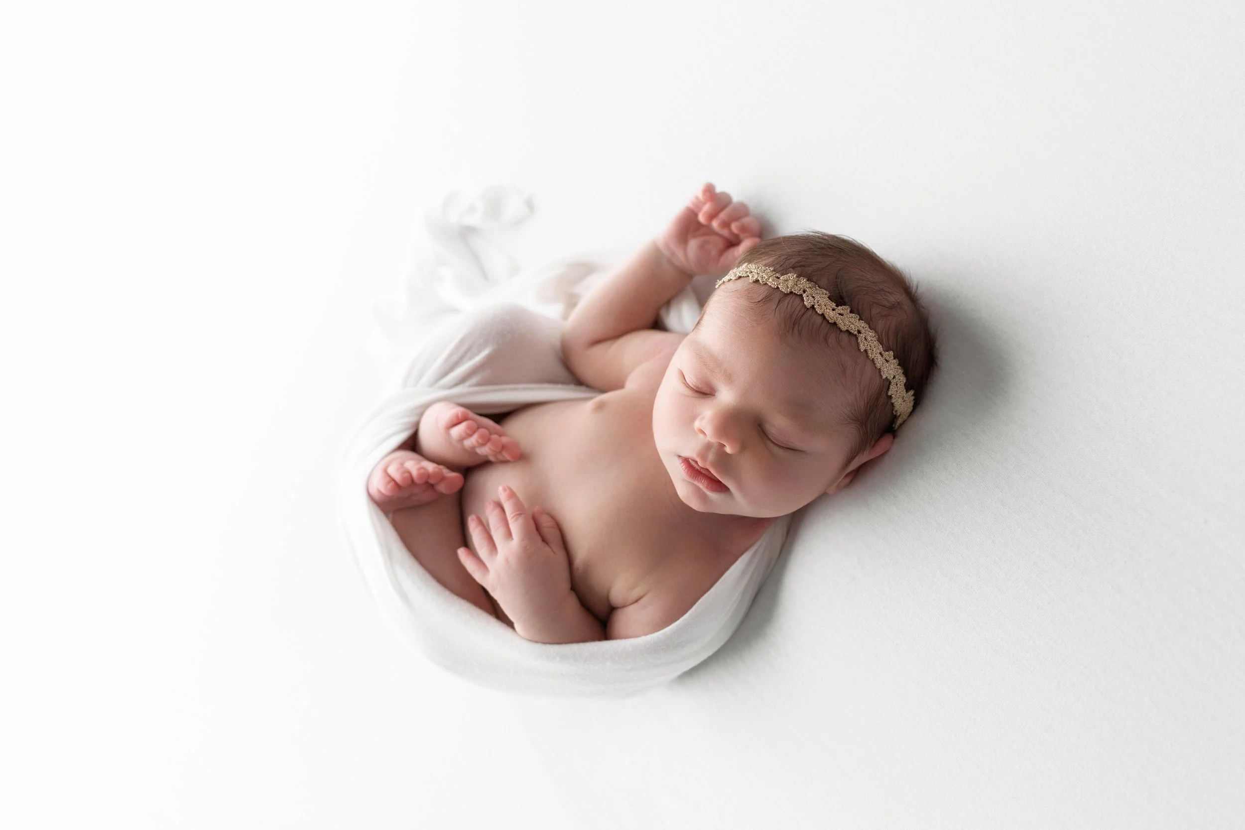 Soft and timeless newborn portrait of a sleeping baby girl wrapped in white fabric with a delicate headband, photographed by a Fayetteville, Arkansas newborn photographer in a clean, neutral studio setting.