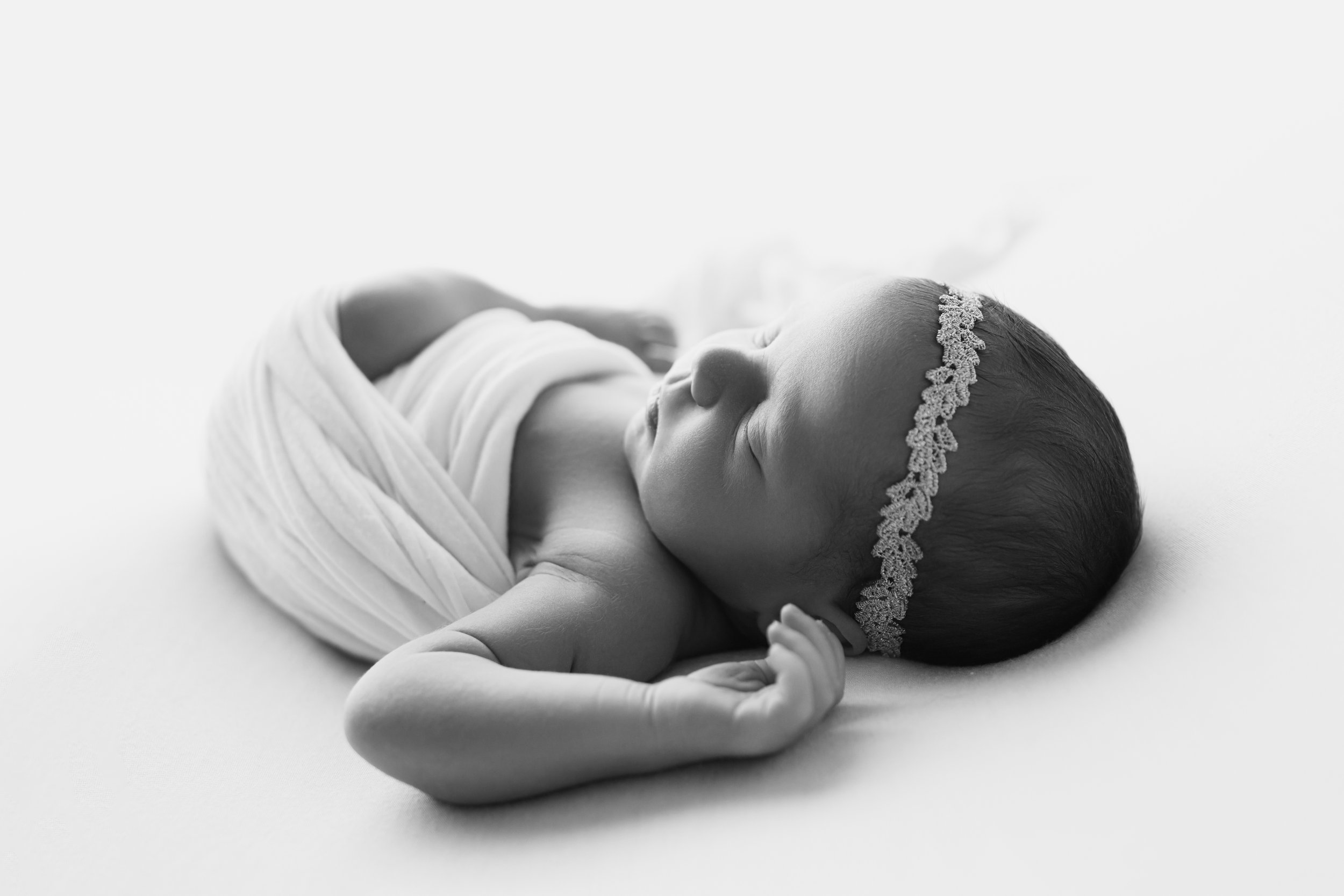 Newborn baby wrapped in a white blanket and wearing a lace headband, sleeping peacefully on a soft surface, located in a Fayetteville, Arkansas studio..