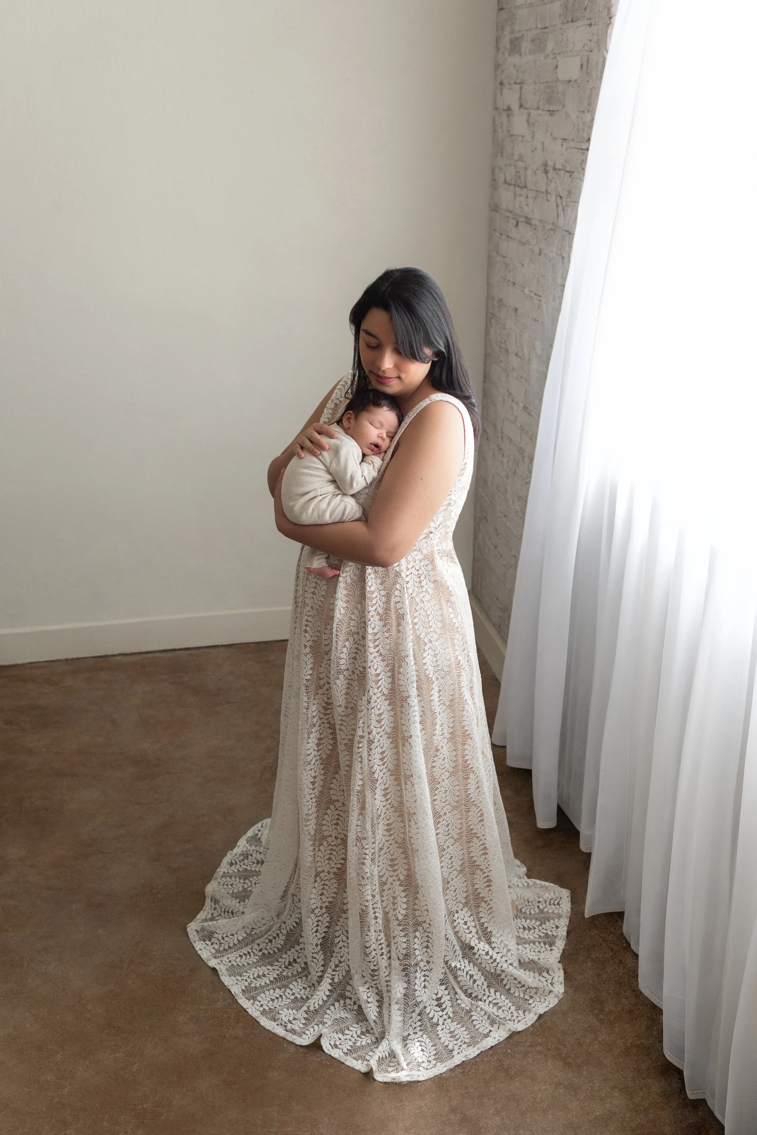 A woman in a long white lace dress holding a sleeping baby in a cream-colored outfit, standing near a white wall with a brick section and a window with white curtains in Fayetteville, Arkansas studio..