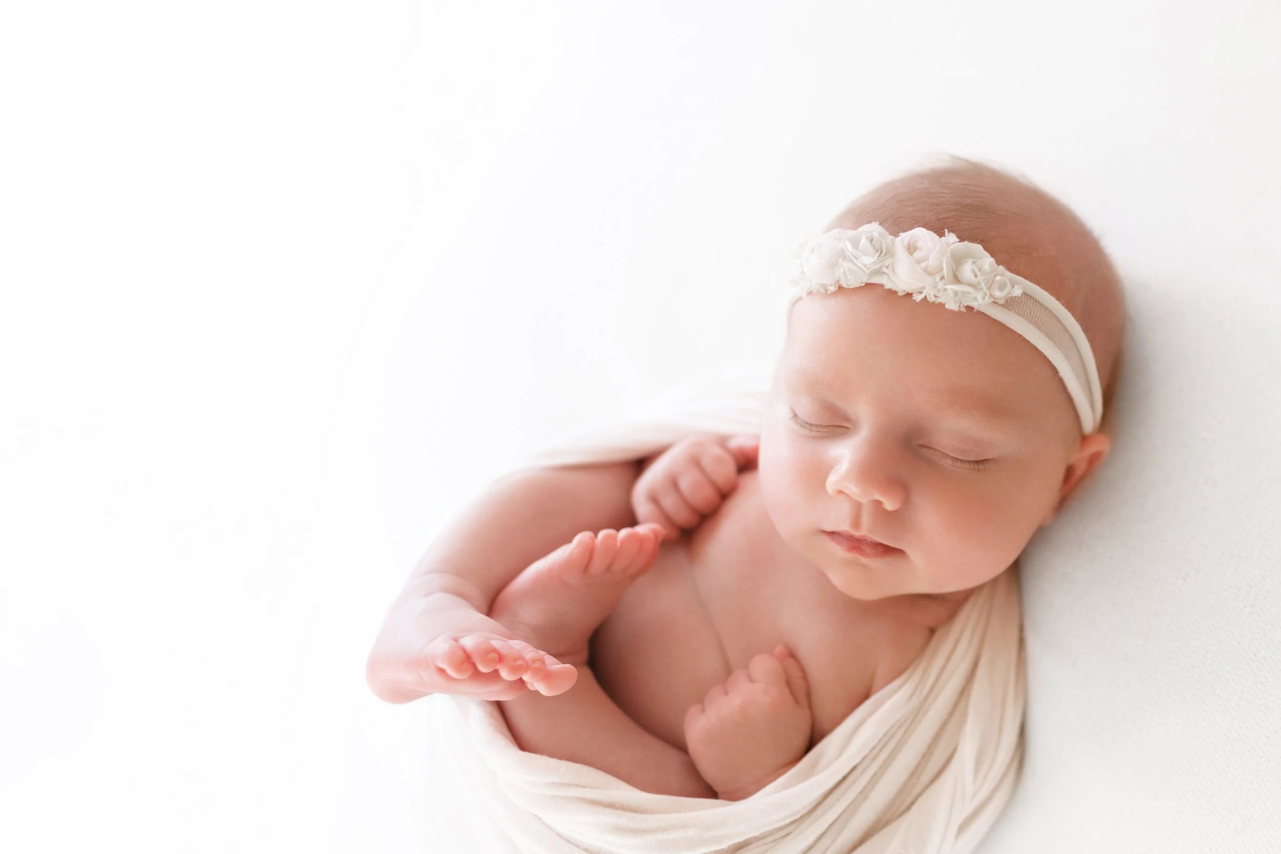 Newborn baby wrapped in a white blanket, wearing a floral headband, sleeping peacefully on a white background in a Fayetteville, Arkansas studio..