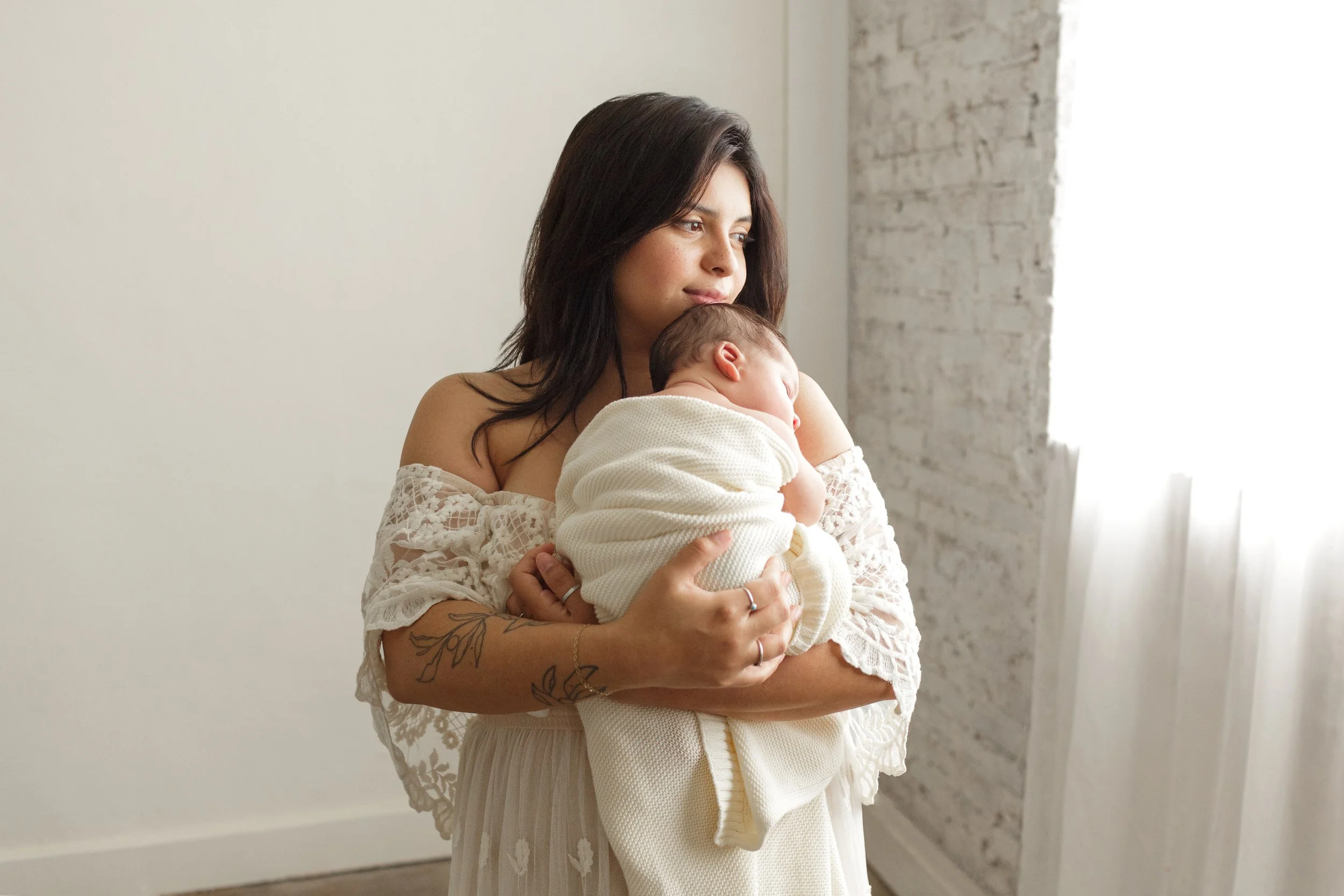 Woman in a white dress holds a swaddled baby close, standing by a bright window in a softly lit room in a Fayetteville, Arkansas studio.