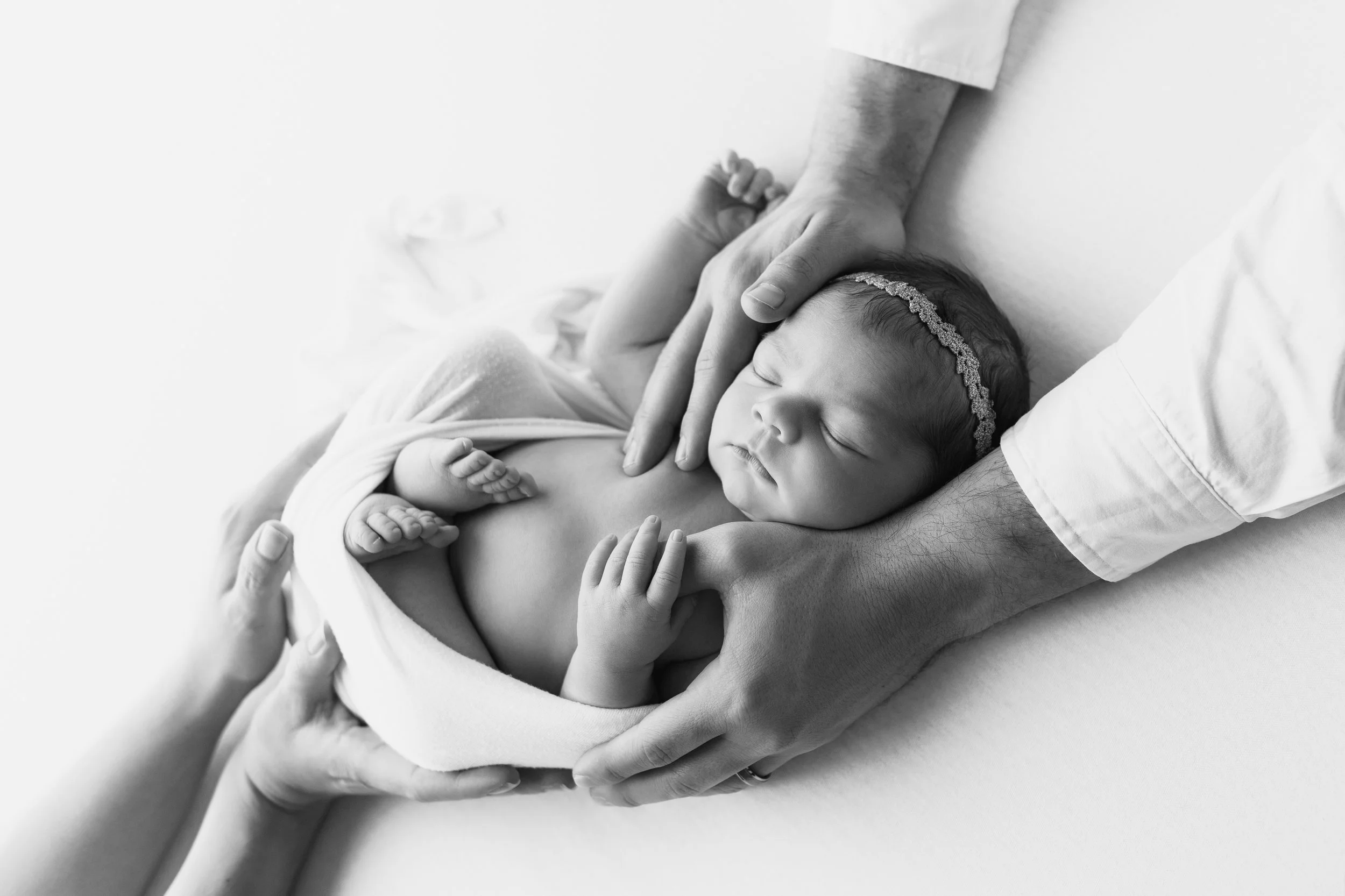 Black and white photo of a newborn baby sleeping, held gently in an adult's hands, wearing a decorative headband.