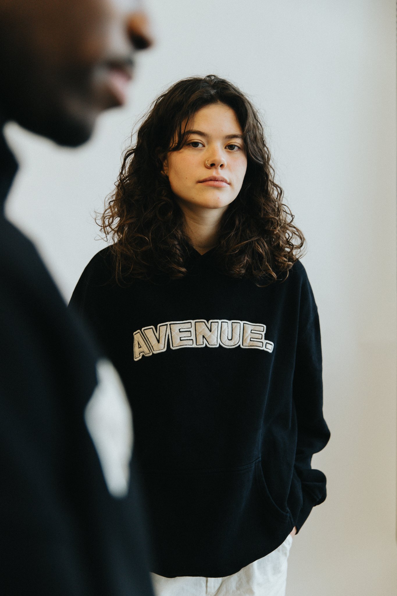 Une jeune femme avec des cheveux bouclés, portant un sweat noir avec l'inscription 'AVENUE' en blanc, regarde vers l'objectif, tandis qu'une autre personne est partiellement visible en premier plan à gauche, floue.