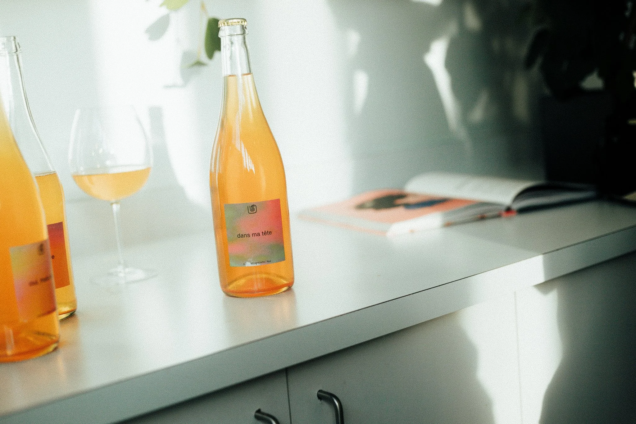 Deux bouteilles d'une boisson orange avec des étiquettes colorées, une verre rempli de la même boisson, un magazine ouvert et une lampe de poche sur une surface blanche, dans un intérieur éclairé par la lumière naturelle.