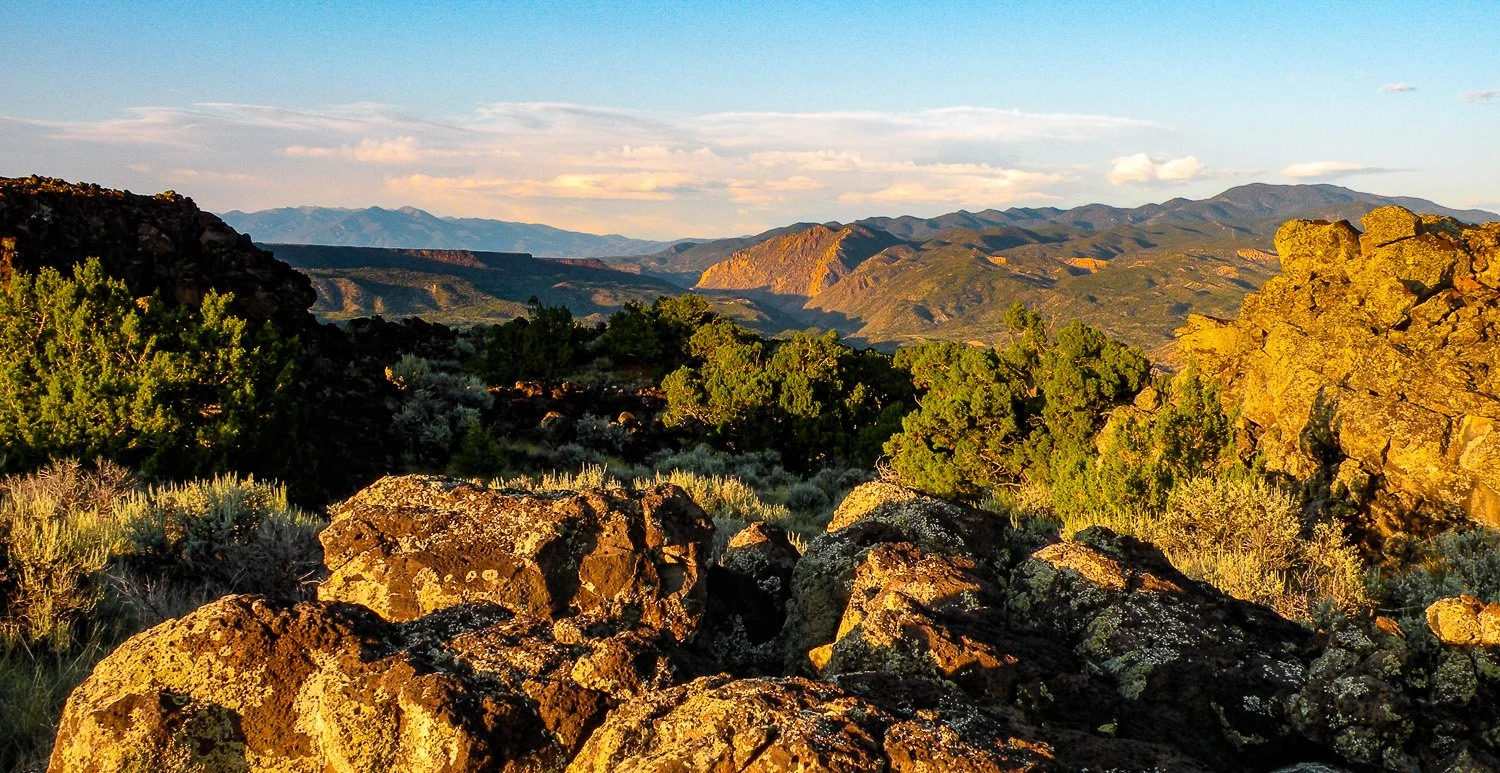 Sunlit rocky landscape with green bushes and distant mountains under a partly cloudy sky.