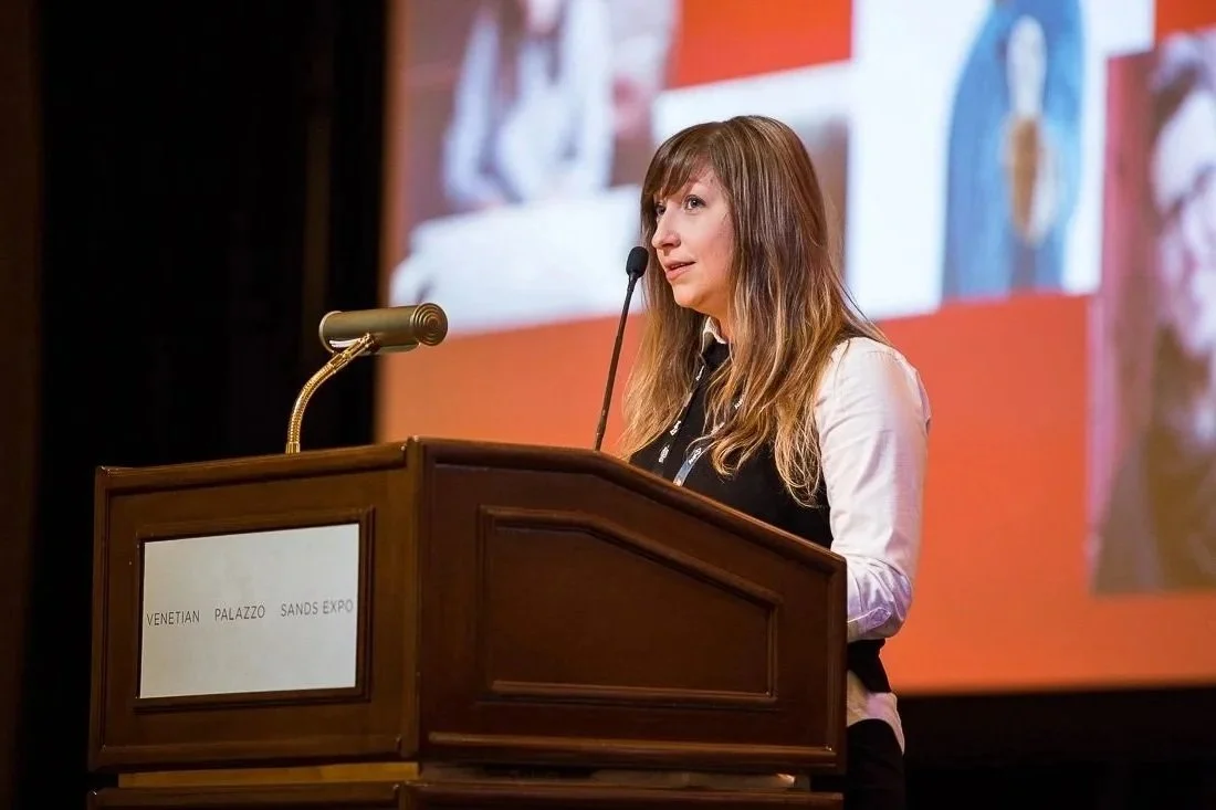 Dr. Jenny is wearing a suit and delivering a keynote standing behind a podium. She has an earnest but hopeful expression.