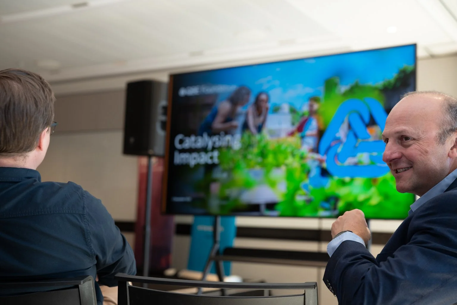 Two men seated in front of a large display screen in an indoor conference room, with one man smiling and talking while the other is looking at the screen. The screen shows people outdoors and text that appears to say "Catalyzing Impact."