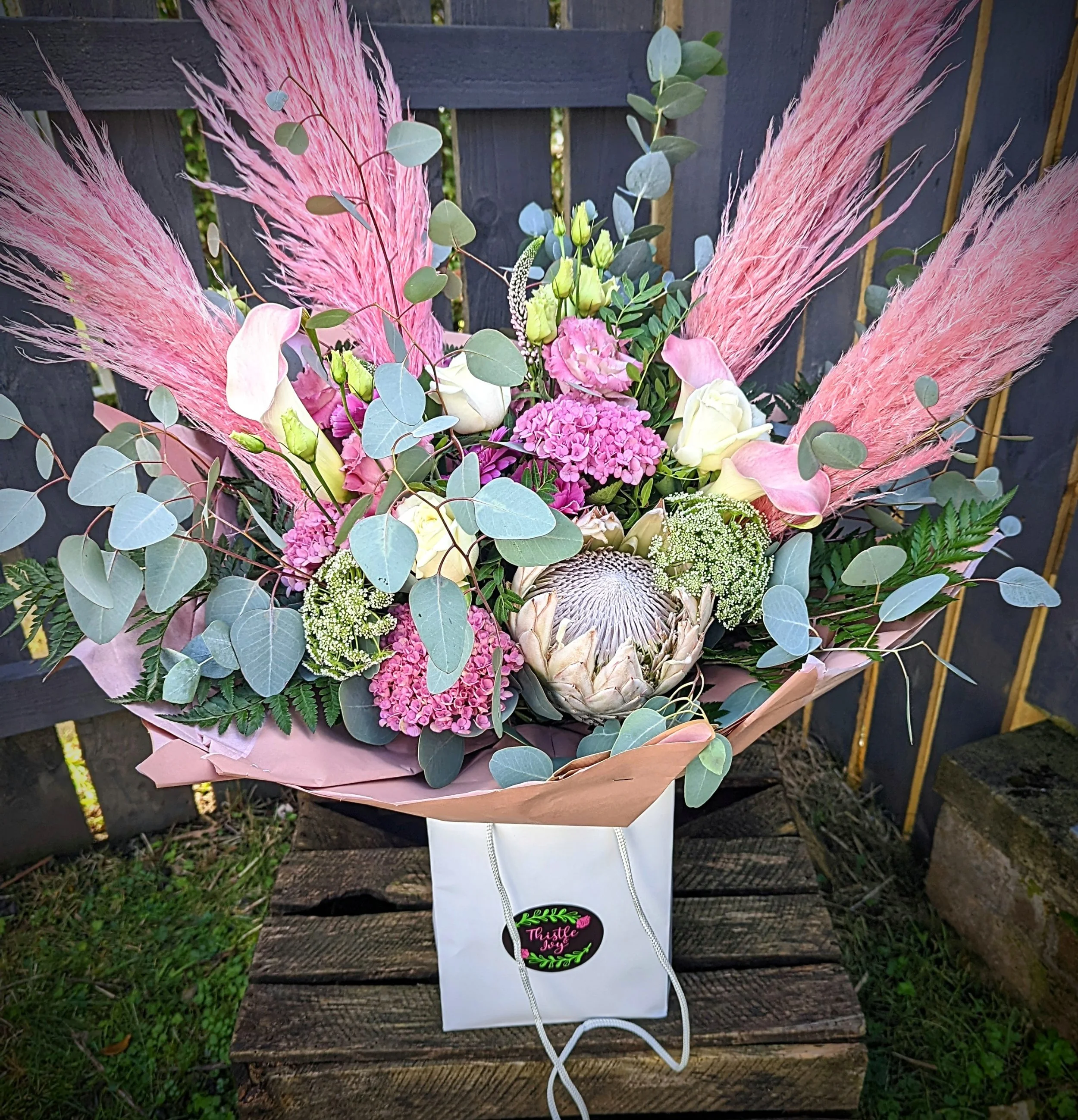 A floral bouquet with pink pampas grass, white roses, pink clarkia, greenery, and a protea flower, in a pink wrapping.