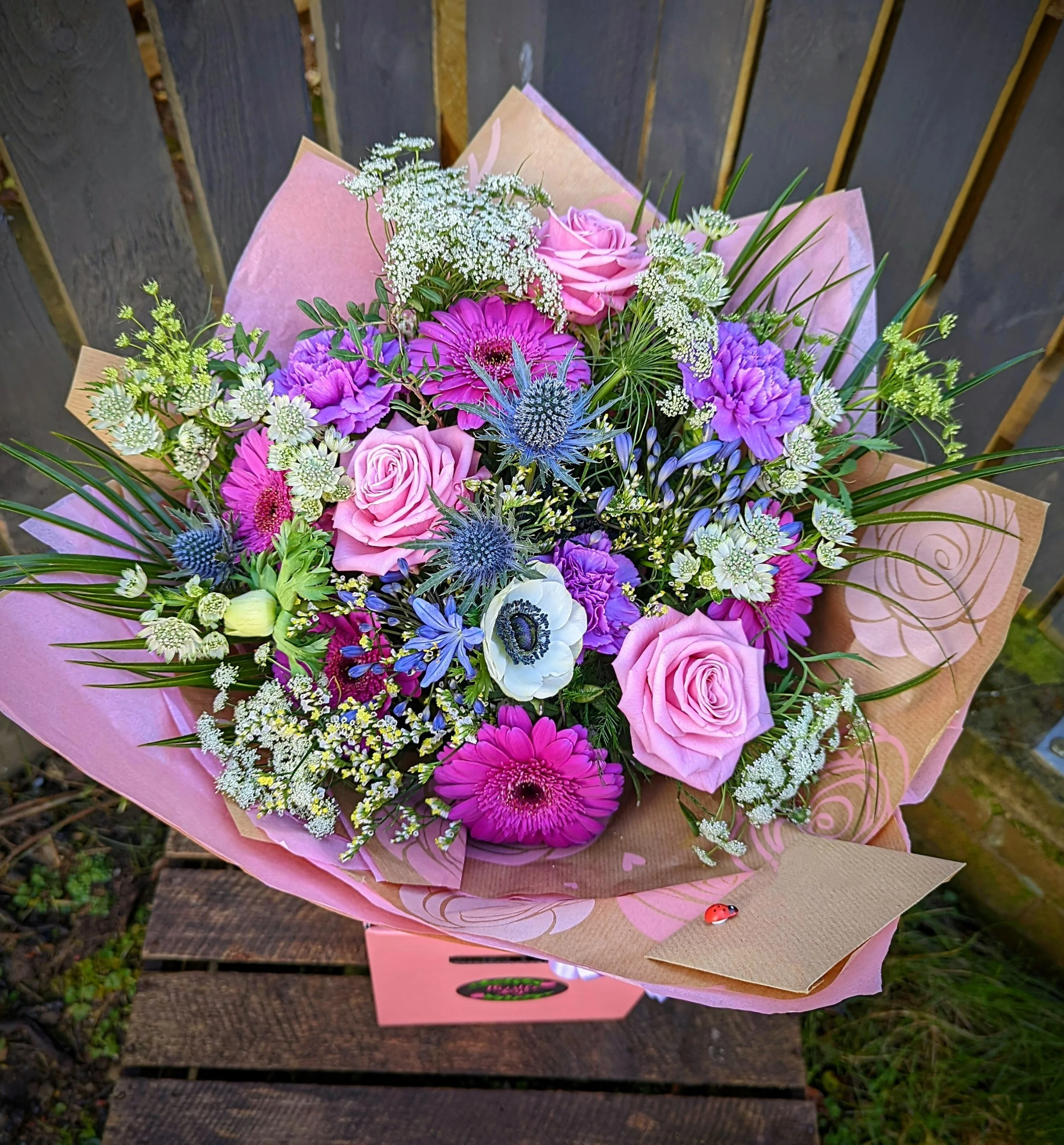 Colorful bouquet of pink, purple, and white flowers with green foliage, wrapped in pink paper, placed on a wooden surface outdoors.