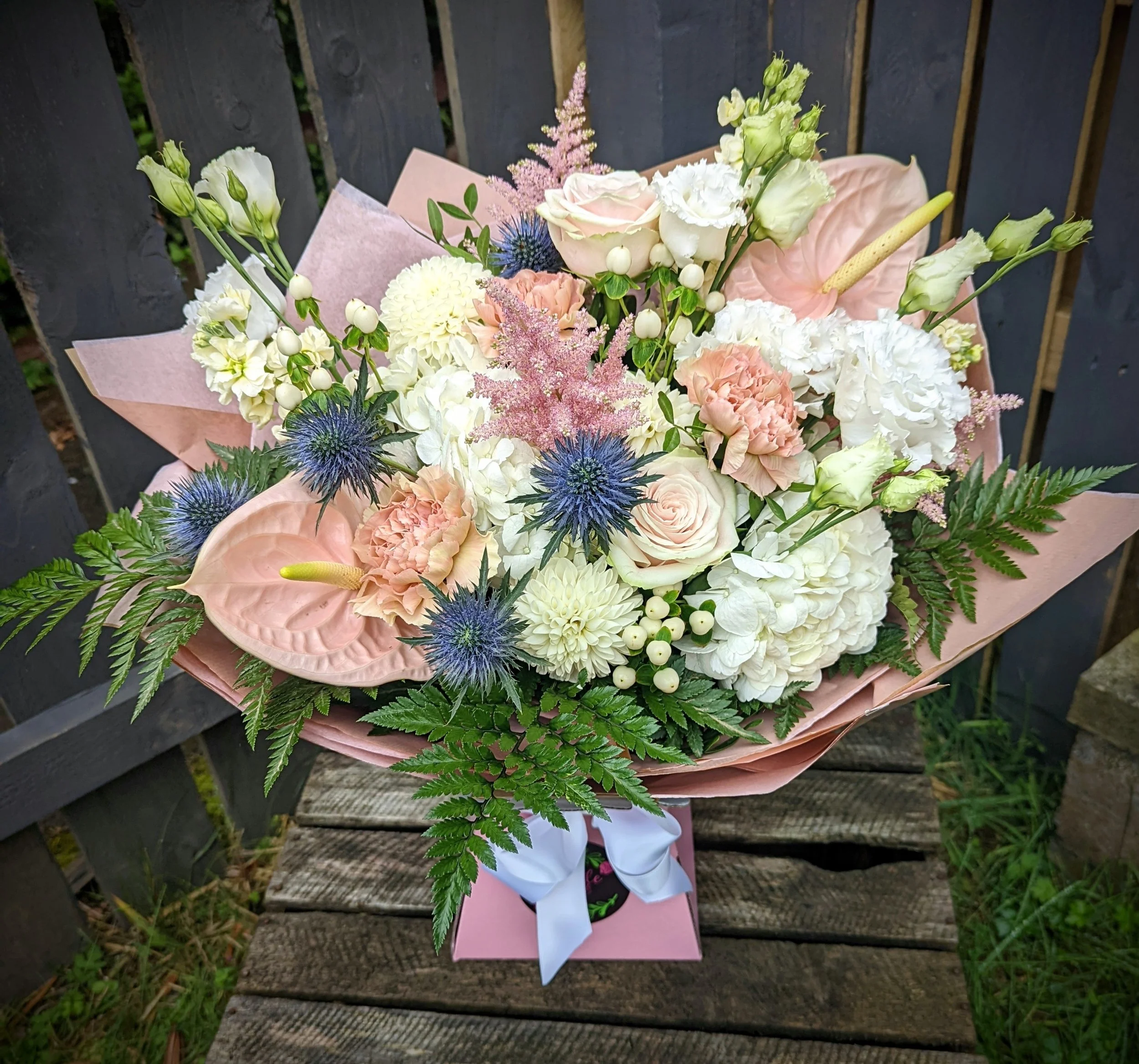 Colorful flower bouquet with roses, hydrangeas, anthuriums, and blue thistle, wrapped in pink paper with a white ribbon, placed on a wooden bench outdoors.