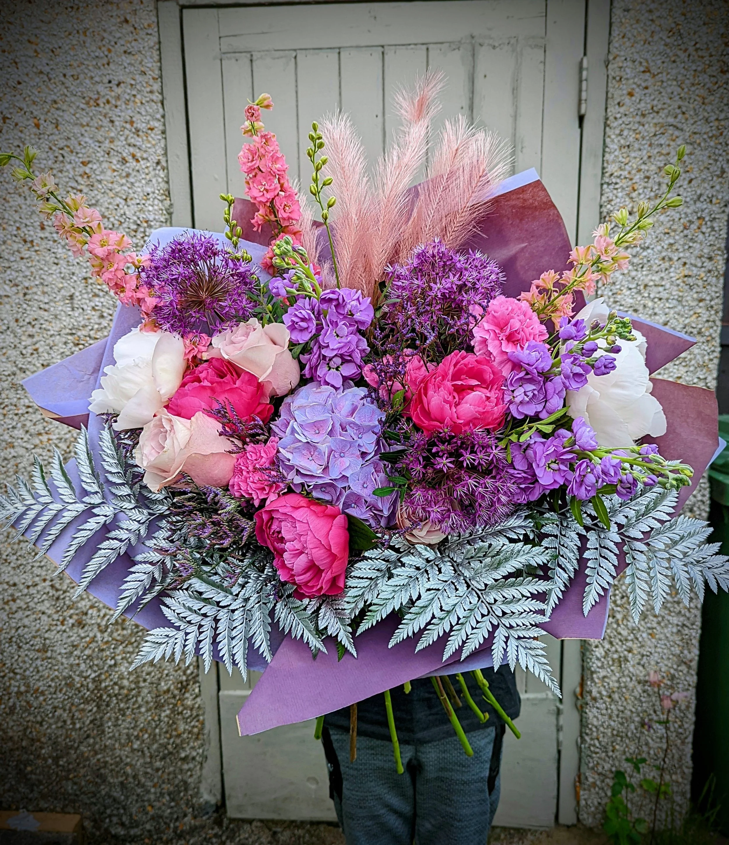 Person holding a large, colorful bouquet of pink, purple, white, and lavender flowers in front of a wooden door.