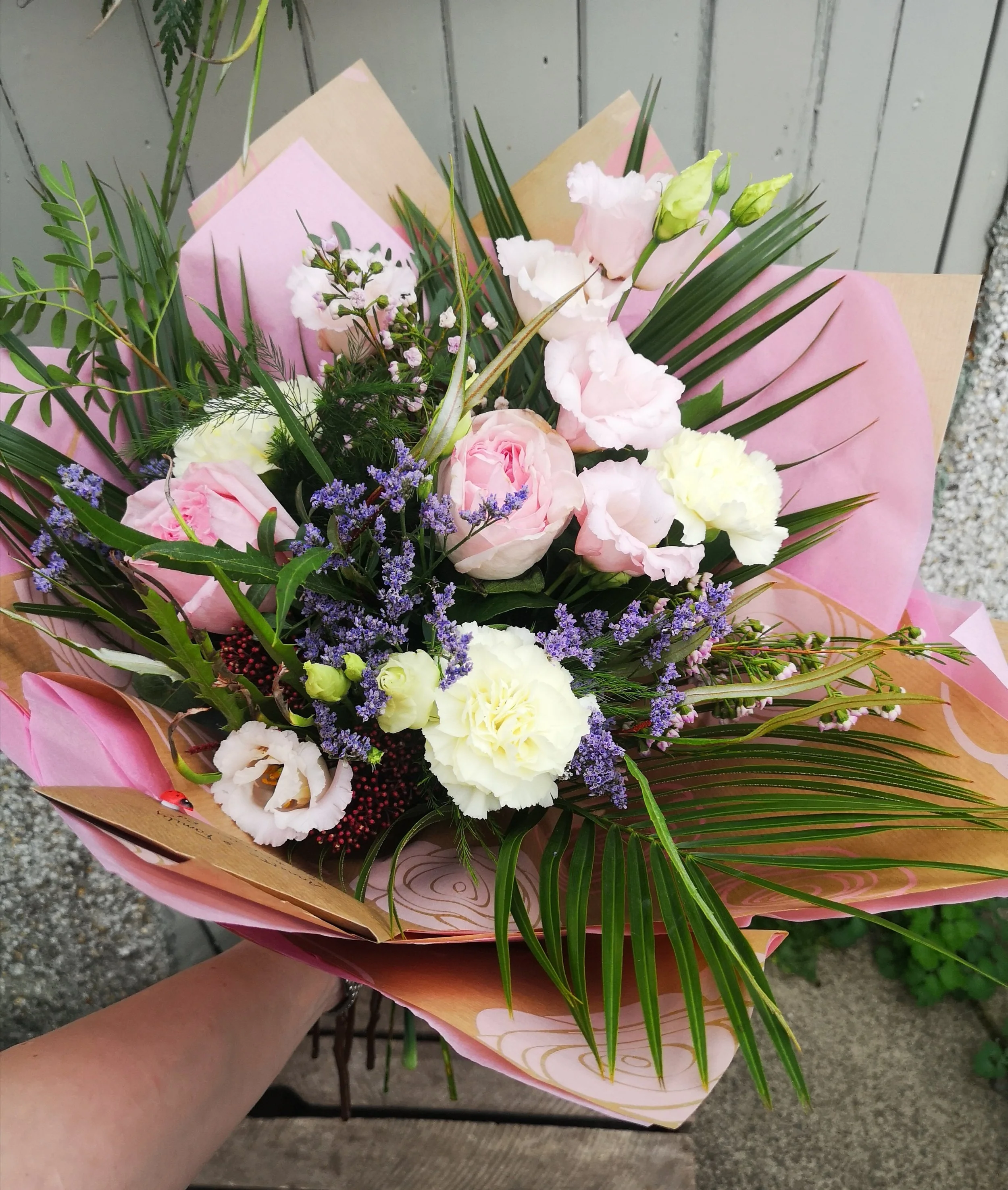 A bouquet of pink, white, and purple flowers wrapped in pink and brown paper with green foliage, held by a person outdoors.