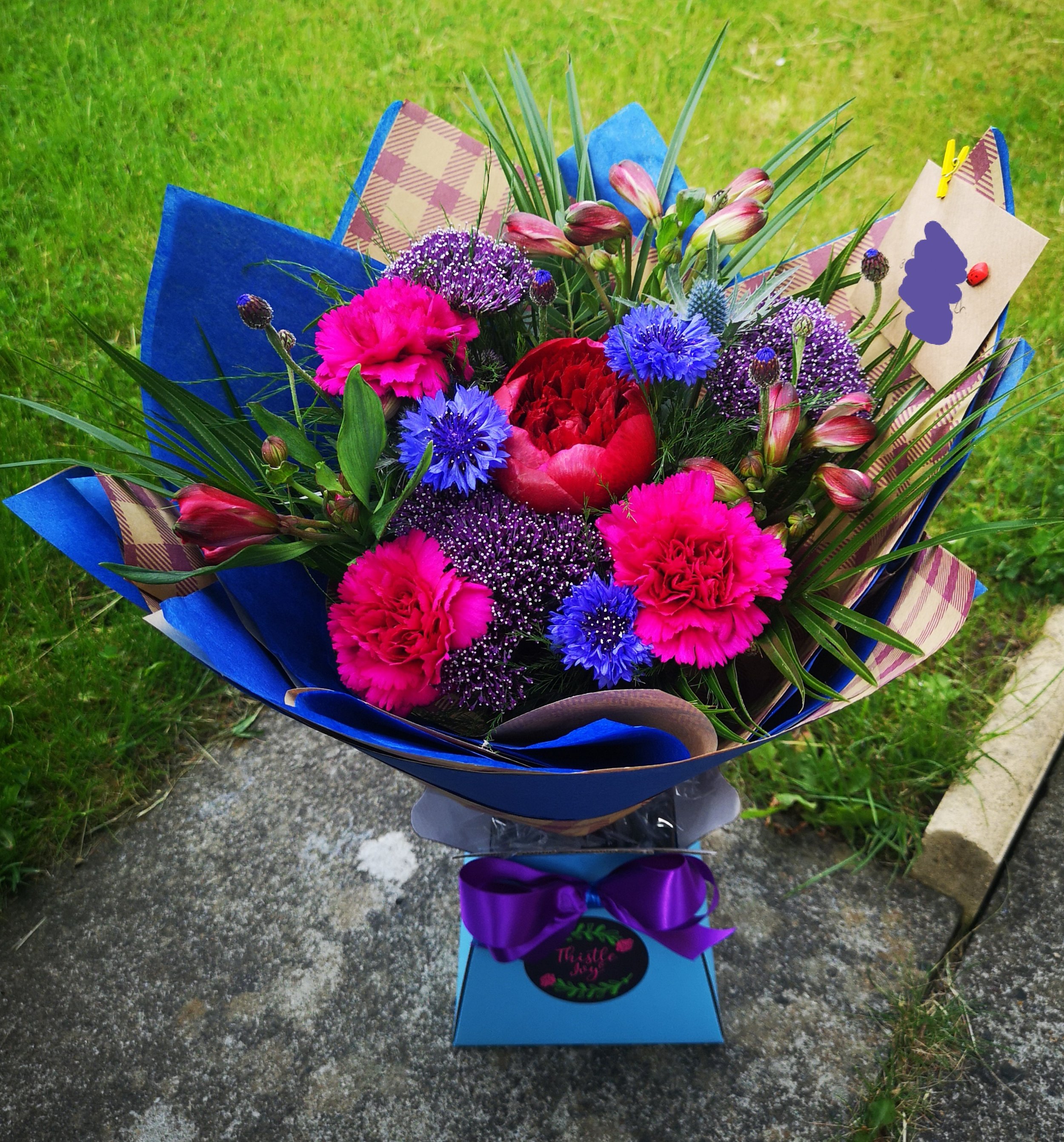 Colorful bouquet of pink, purple, and blue flowers arranged in a blue wrapping with a purple ribbon, placed on a concrete surface with grass in the background.