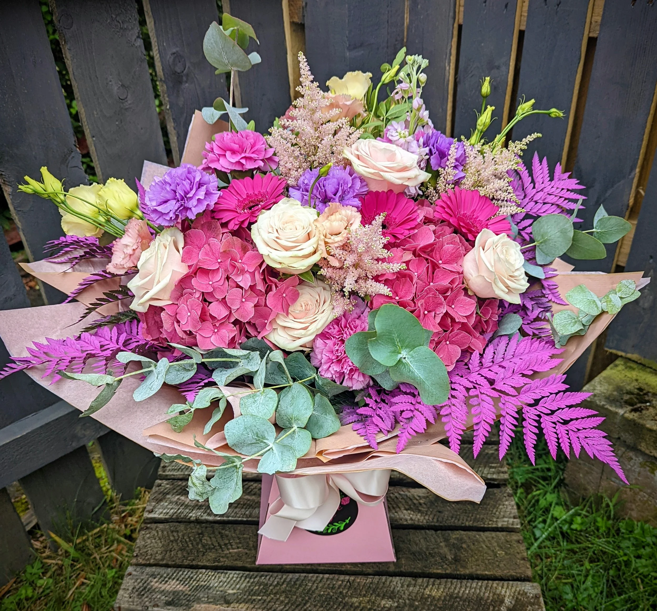 A colorful bouquet of pink, purple, and cream flowers, including roses, hydrangeas, and carnations, accented with green and purple foliage, wrapped in pink paper with a white ribbon, placed on a wooden surface outdoors.