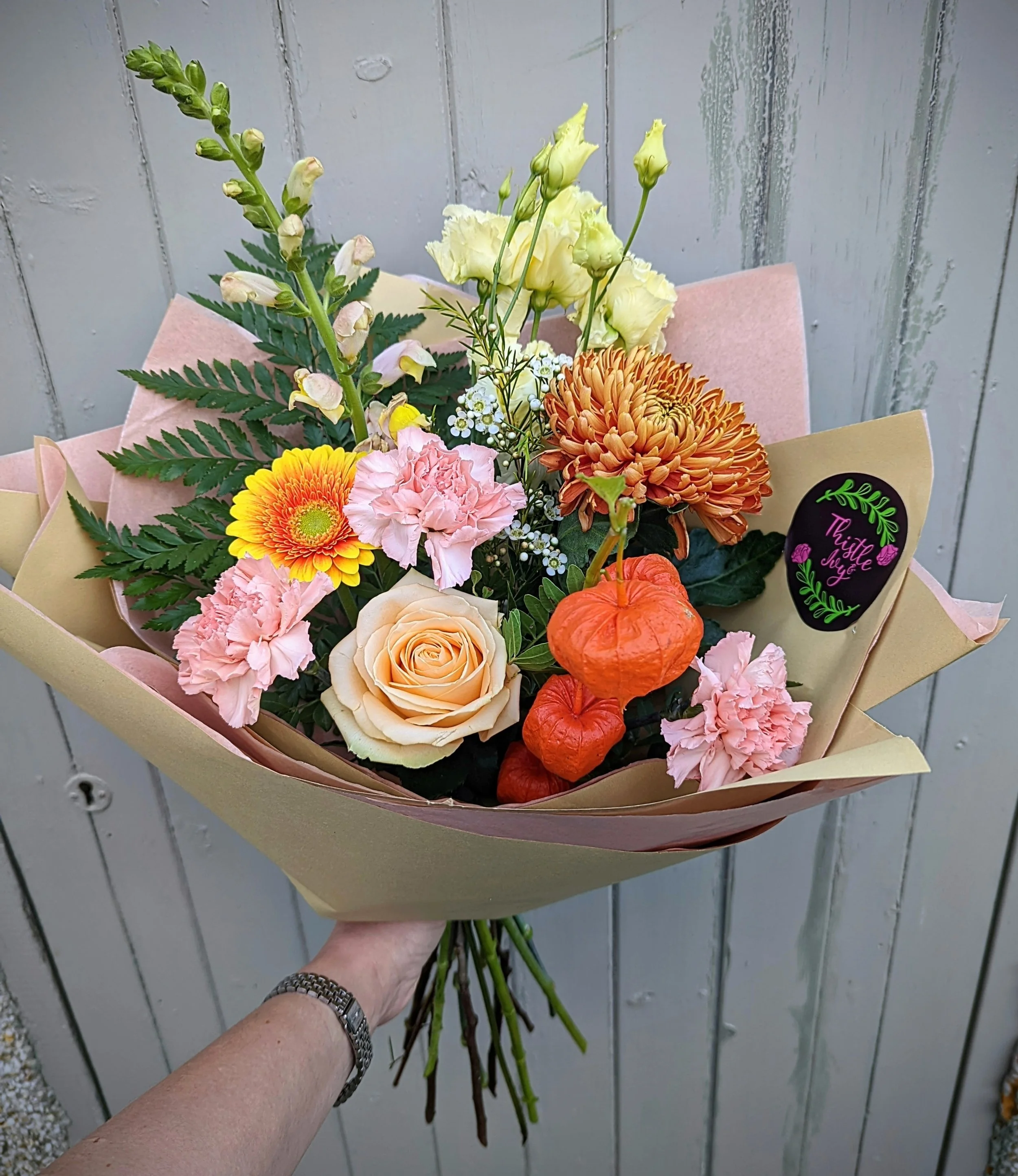 A bouquet of various colorful flowers including roses, chrysanthemums, and lilies, wrapped in beige and pink paper, held by a person wearing a watch.