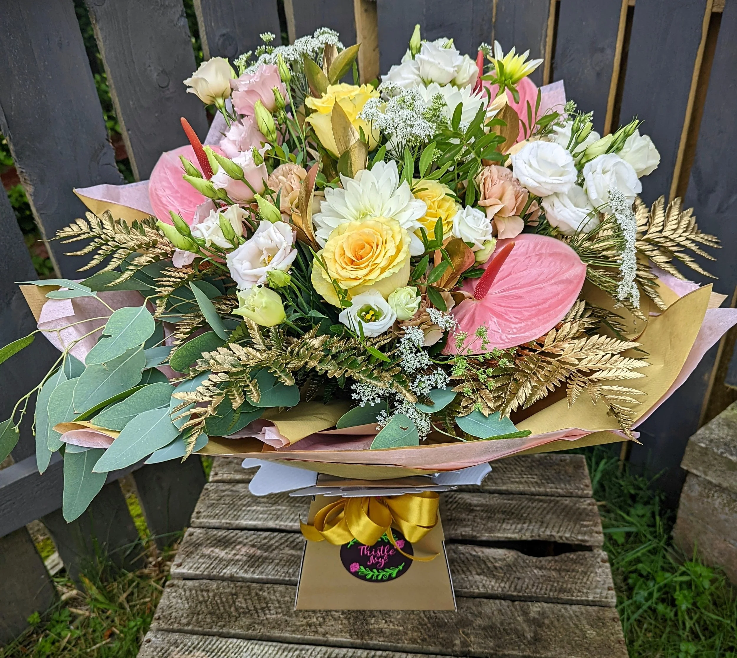 Colorful bouquet of flowers with pink anthuriums, white roses, yellow roses, white daisies, and various greenery, wrapped in pink and gold paper, and placed in a gift box with a gold ribbon.
