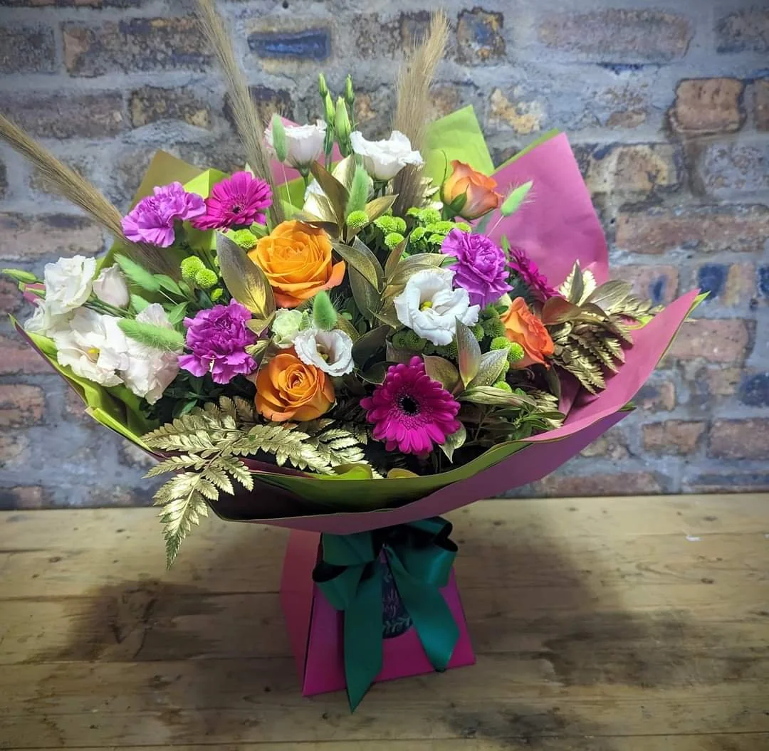 Colorful bouquet of flowers including roses, gerbera daisies, lisianthus, and greenery in pink wrapping paper with a green and black ribbon.